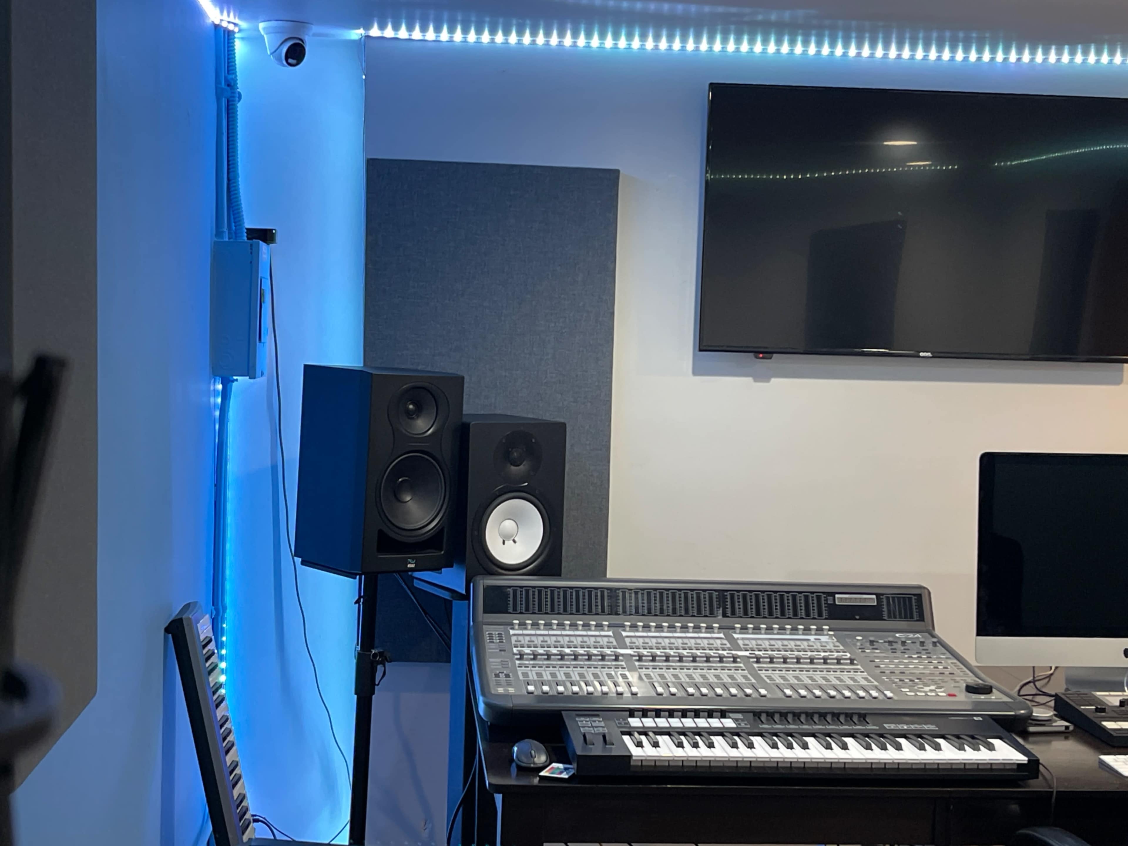 The image shows a recording studio featuring a digital audio workstation, a sound mixing console, studio monitors on stands, and a television mounted on the wall.