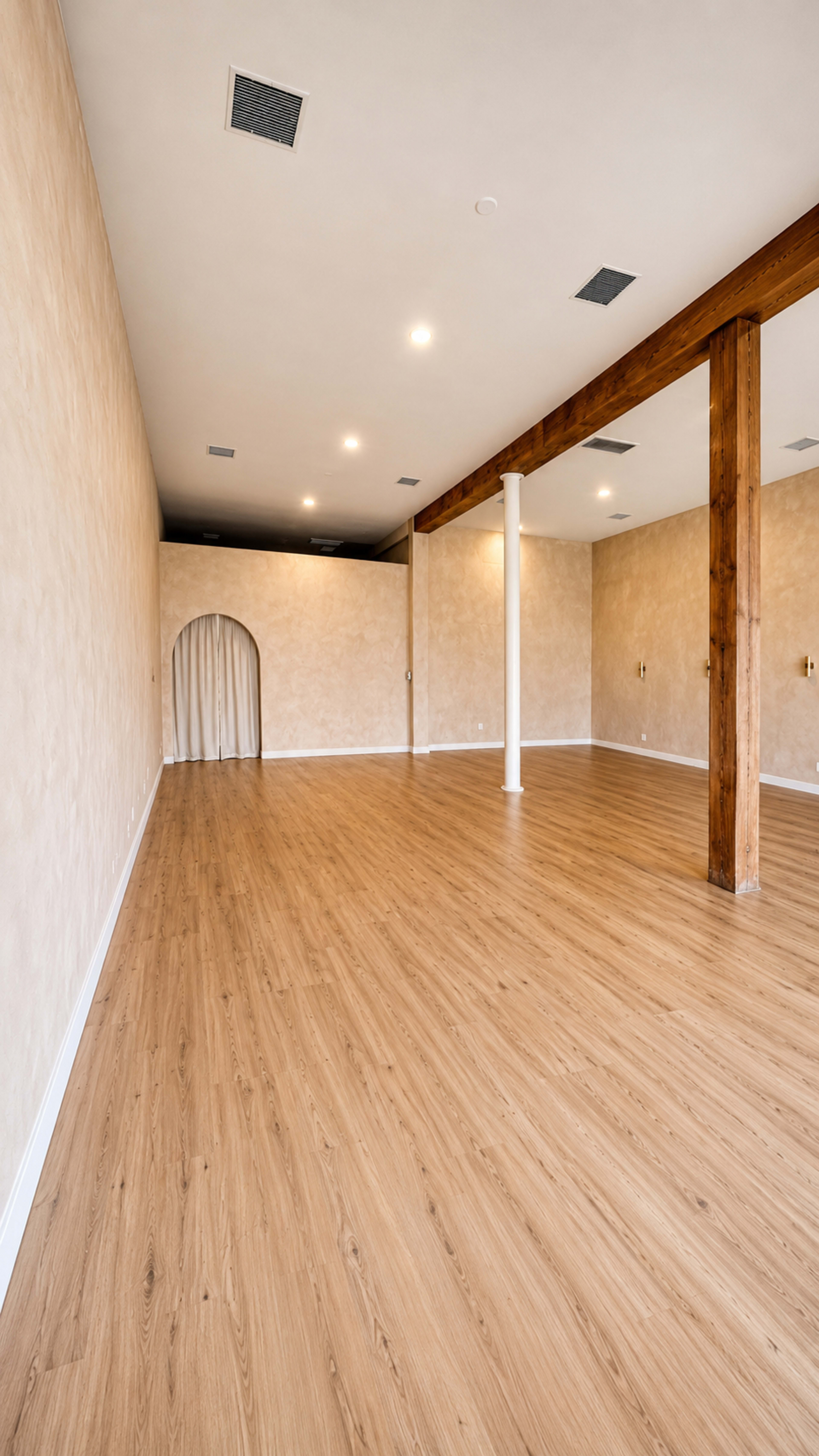 The image shows a spacious, empty room with wooden flooring and pale walls, featuring a decorative archway and wooden support beams.
