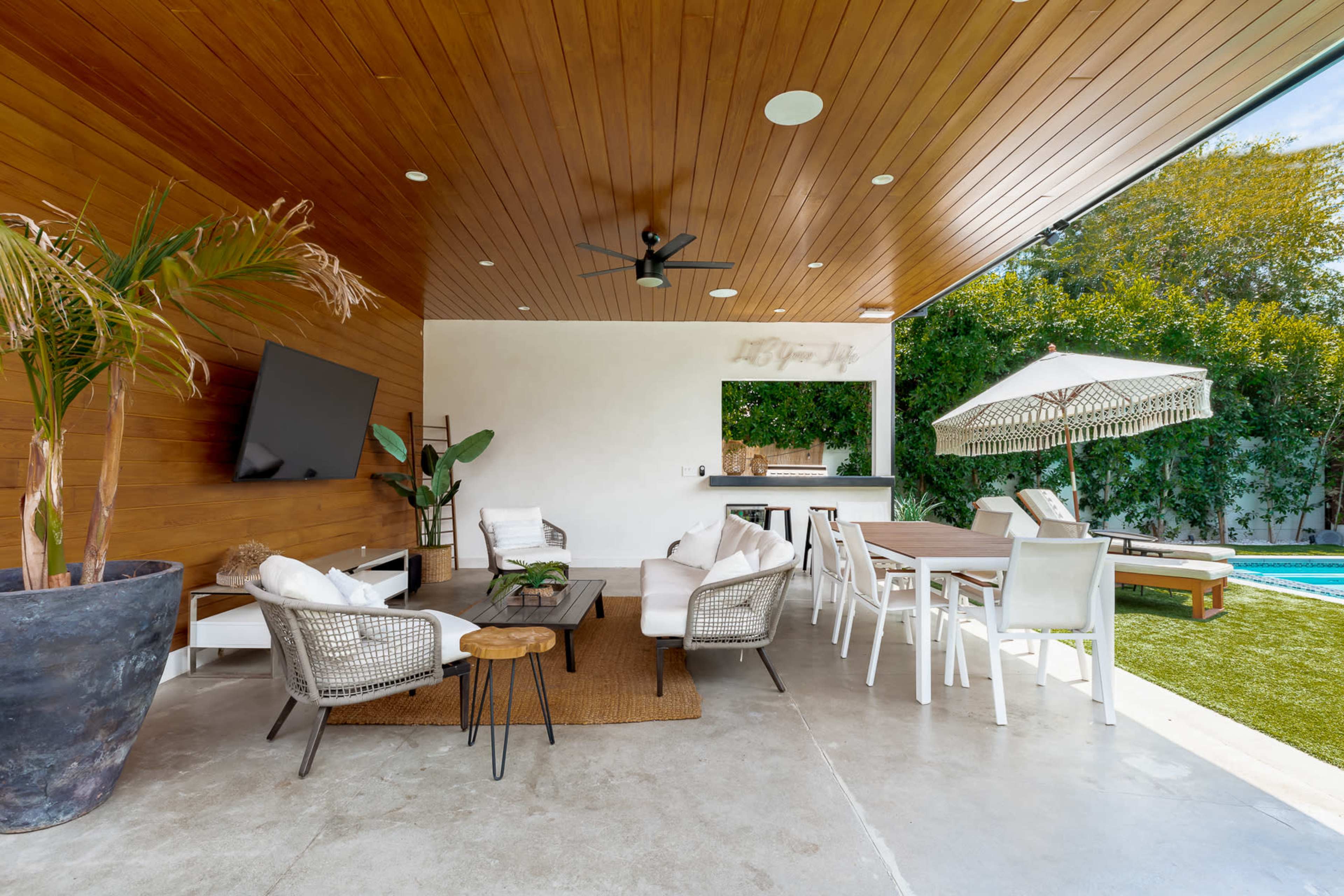 The image shows a contemporary outdoor living space with seating, a dining table, and a pool visible in the background.