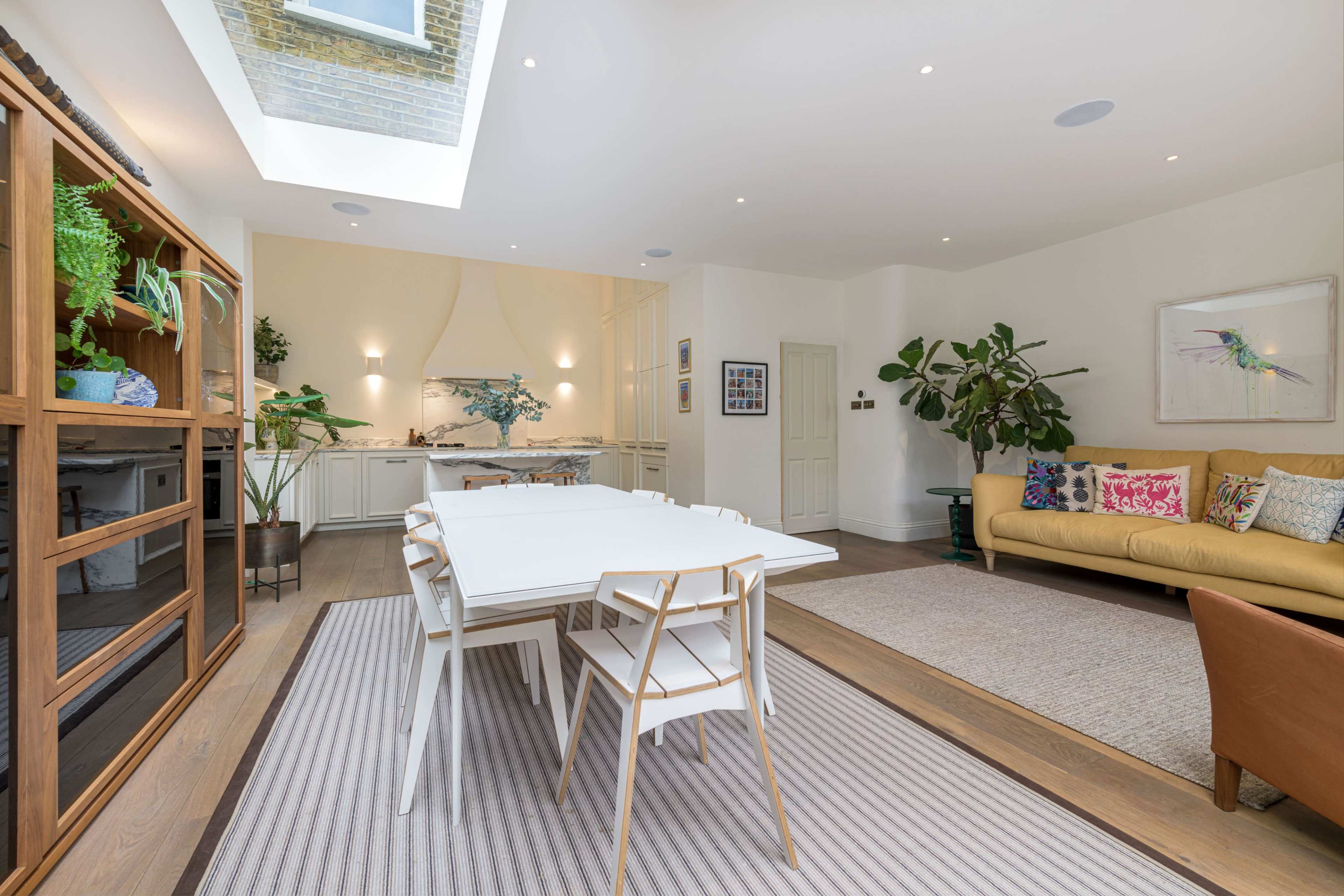 A modern, brightly lit living space featuring a dining area with a white table surrounded by chairs, a couch, plants, and an open kitchen in the background.