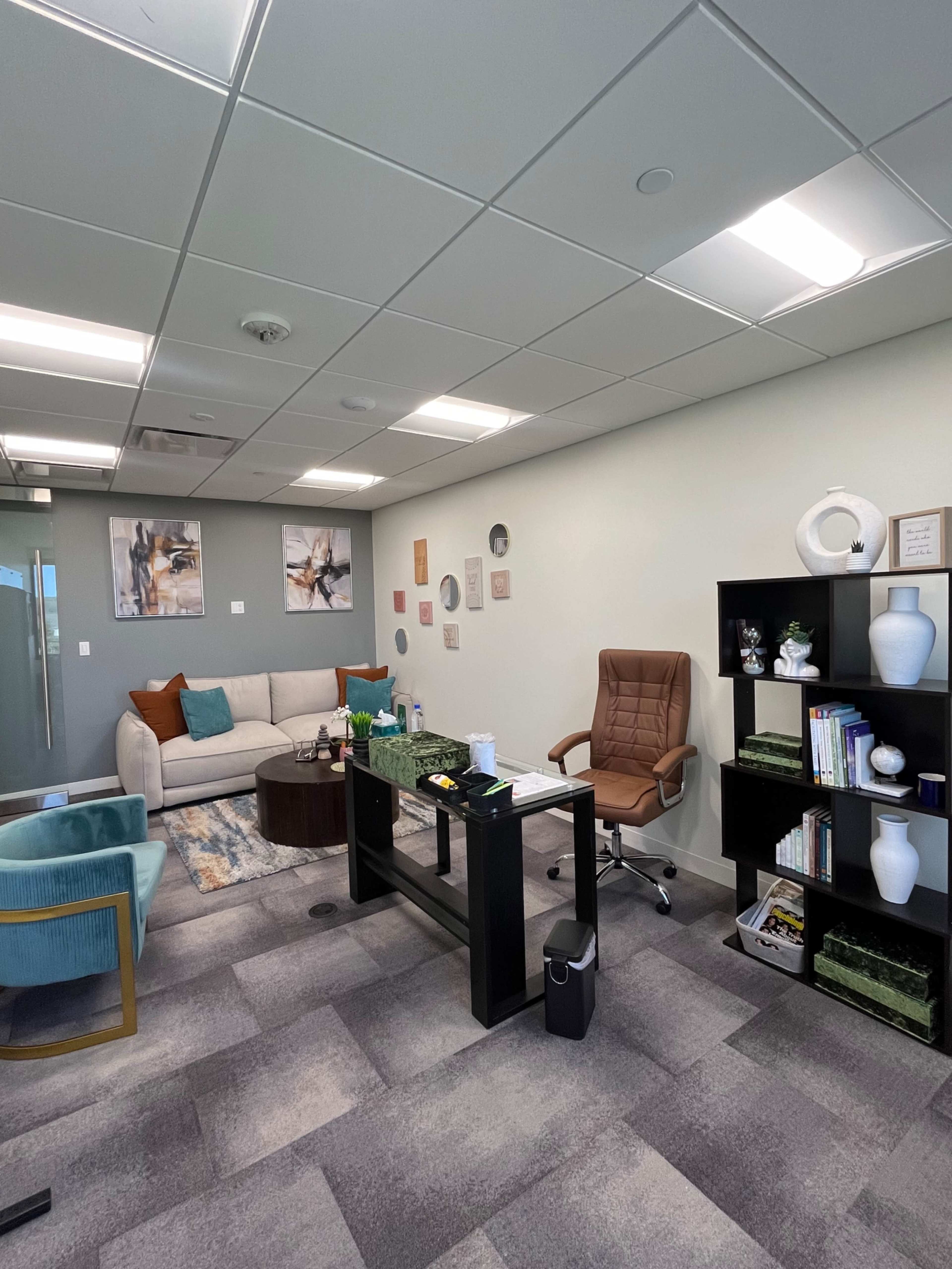A modern office space featuring a beige sofa, a wooden coffee table, an ergonomic chair at a desk, and shelving with various decorative items and books.