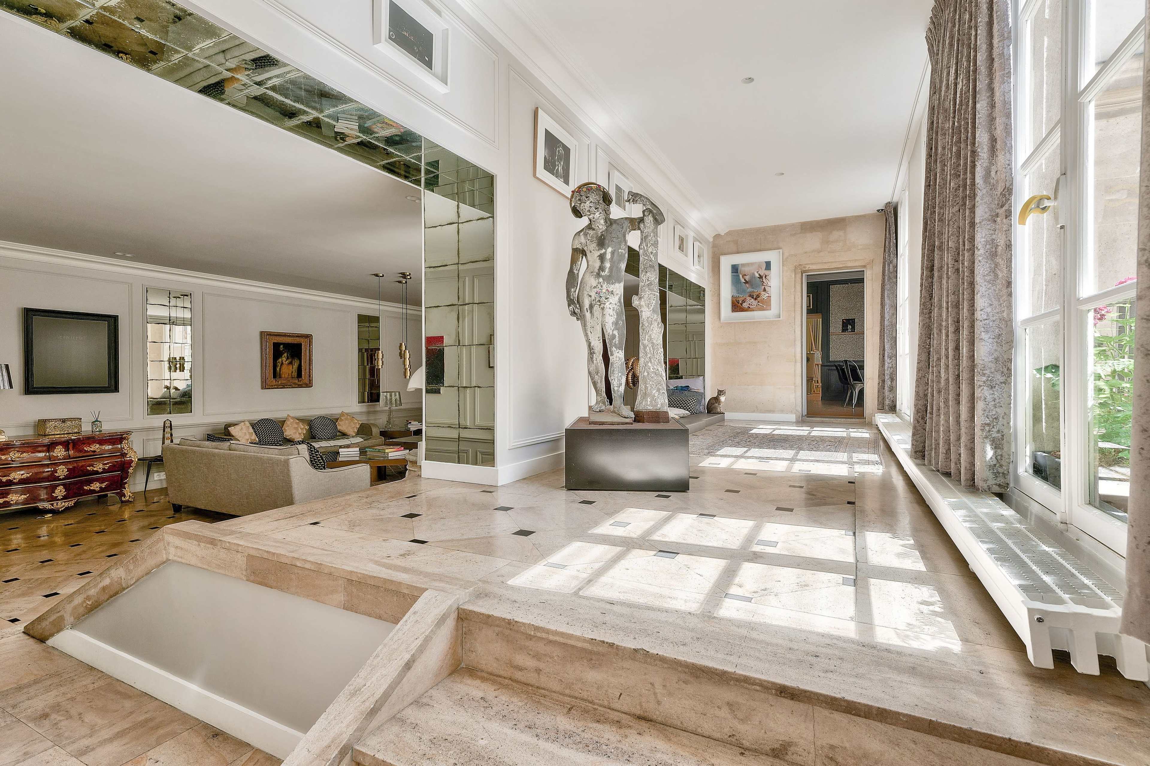 The image shows a spacious, elegantly designed living area with large windows allowing natural light, a mix of contemporary art and furnishings, and a sculpture prominently displayed on a step-up platform.