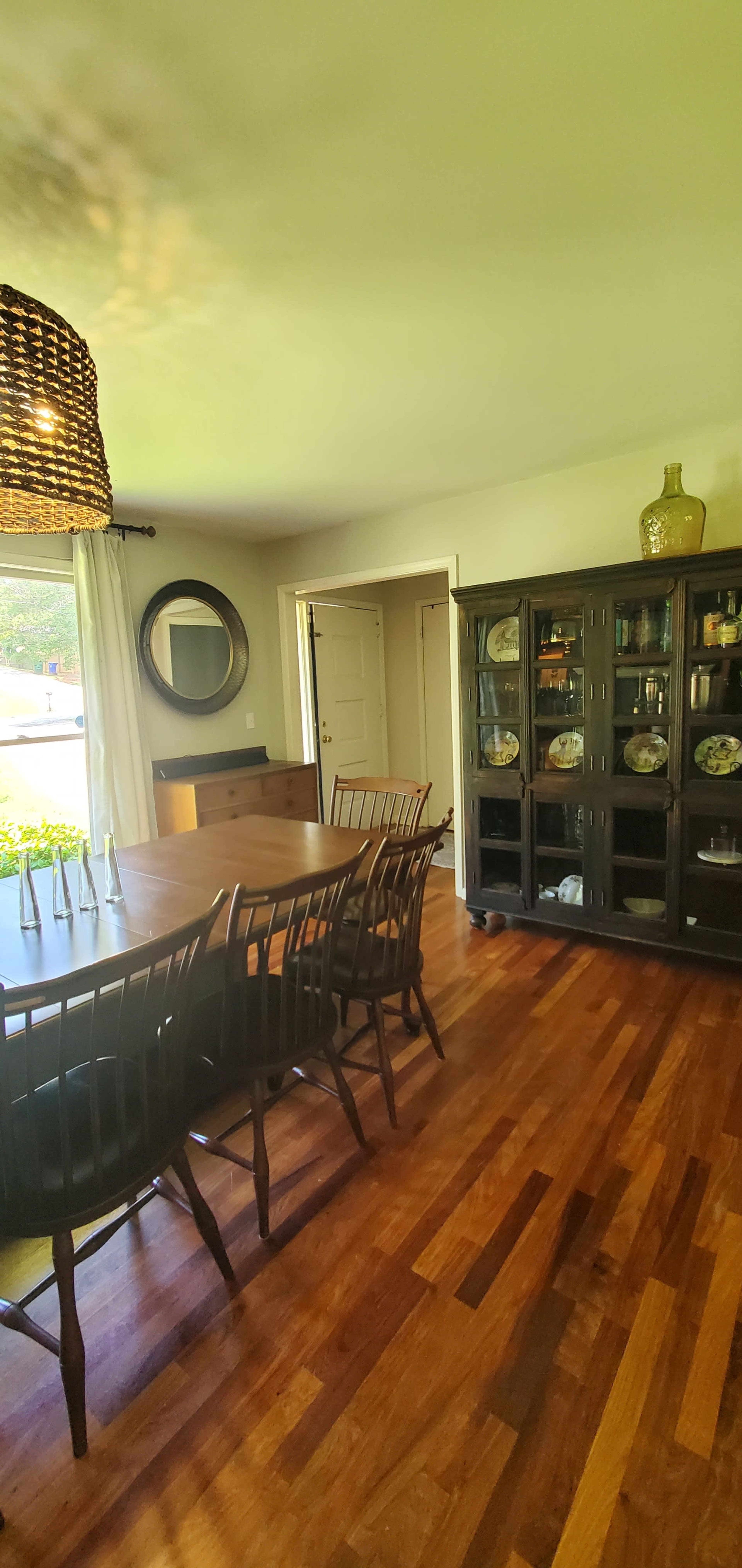 A dining room features a long wooden table with seating around it, a large glass-front cabinet filled with decor, and a circular mirror on the wall.