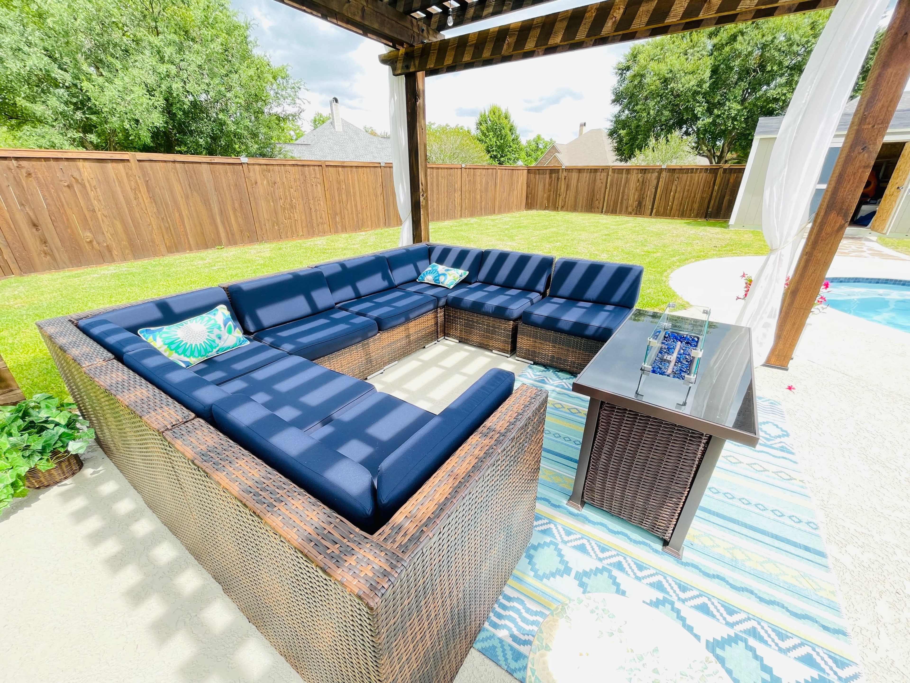 A spacious outdoor seating area features a dark blue sectional sofa arranged around a glass-top table, set on a patterned rug beside a swimming pool.
