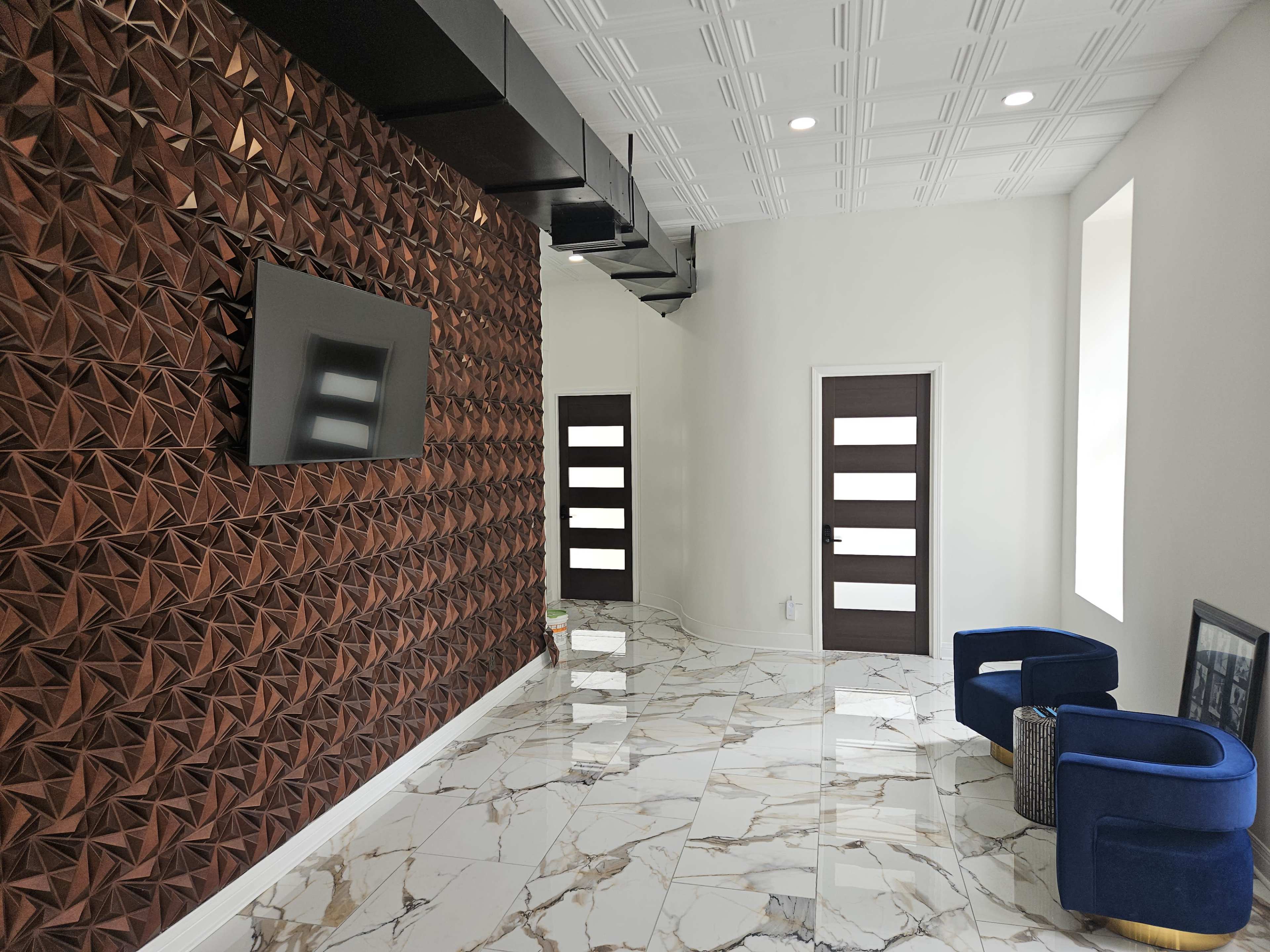 The room features a geometric patterned wall, a television, marble flooring, and modern blue armchairs beside a door with vertical panes.