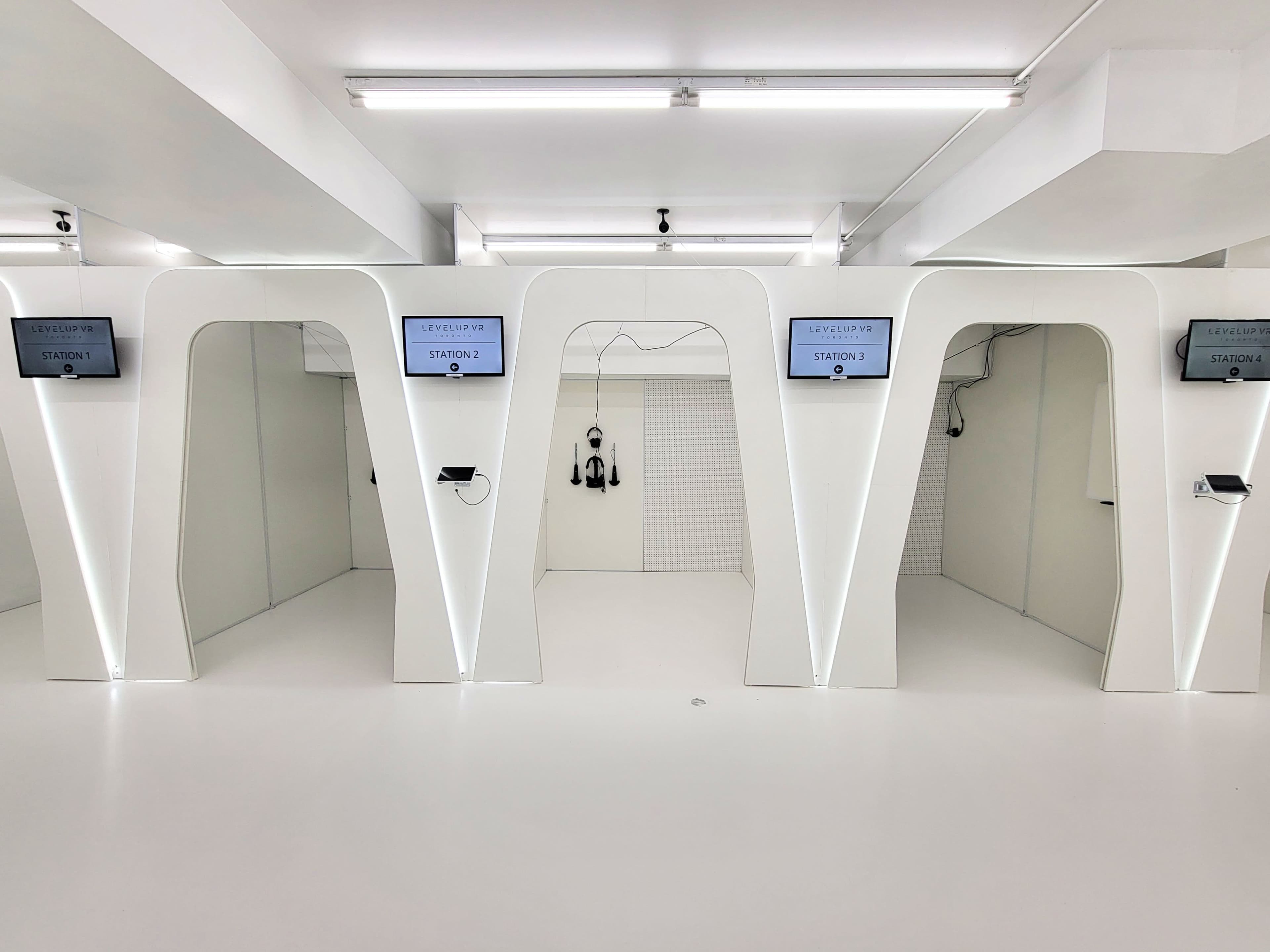 The image shows a modern, minimalist room featuring several white, arch-shaped stations with screens and equipment, set against a white background.