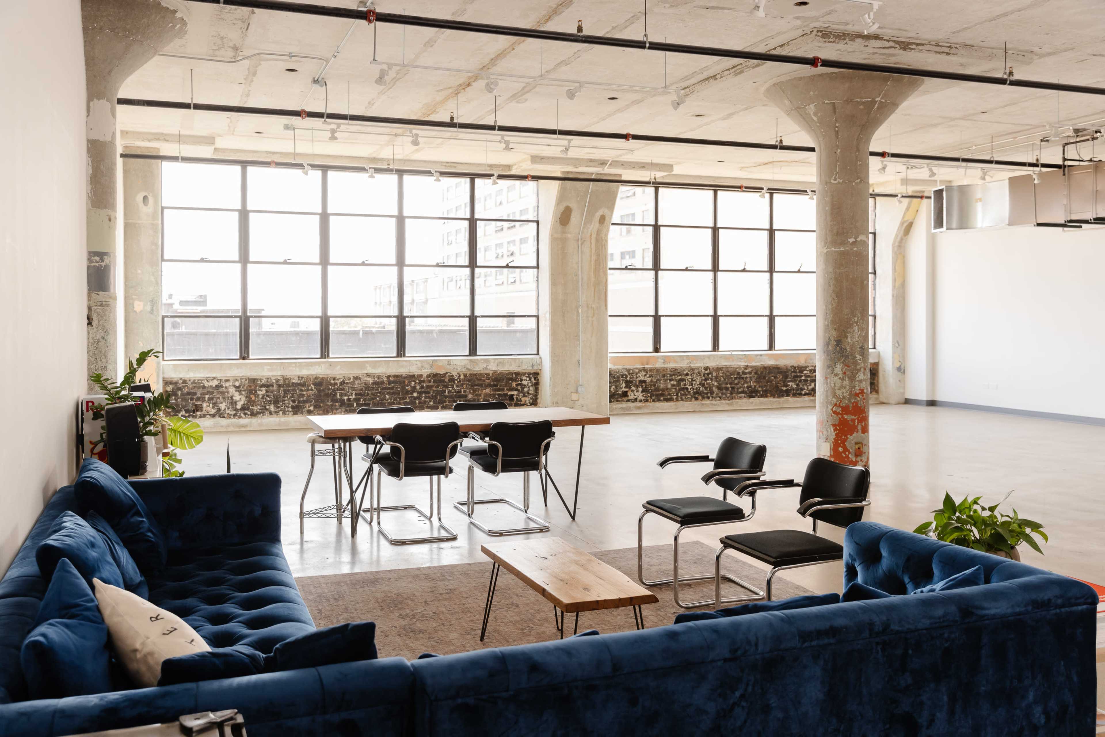 A spacious, industrial-style room with large windows, a blue velvet sofa, a wooden table surrounded by chairs, and exposed concrete pillars.