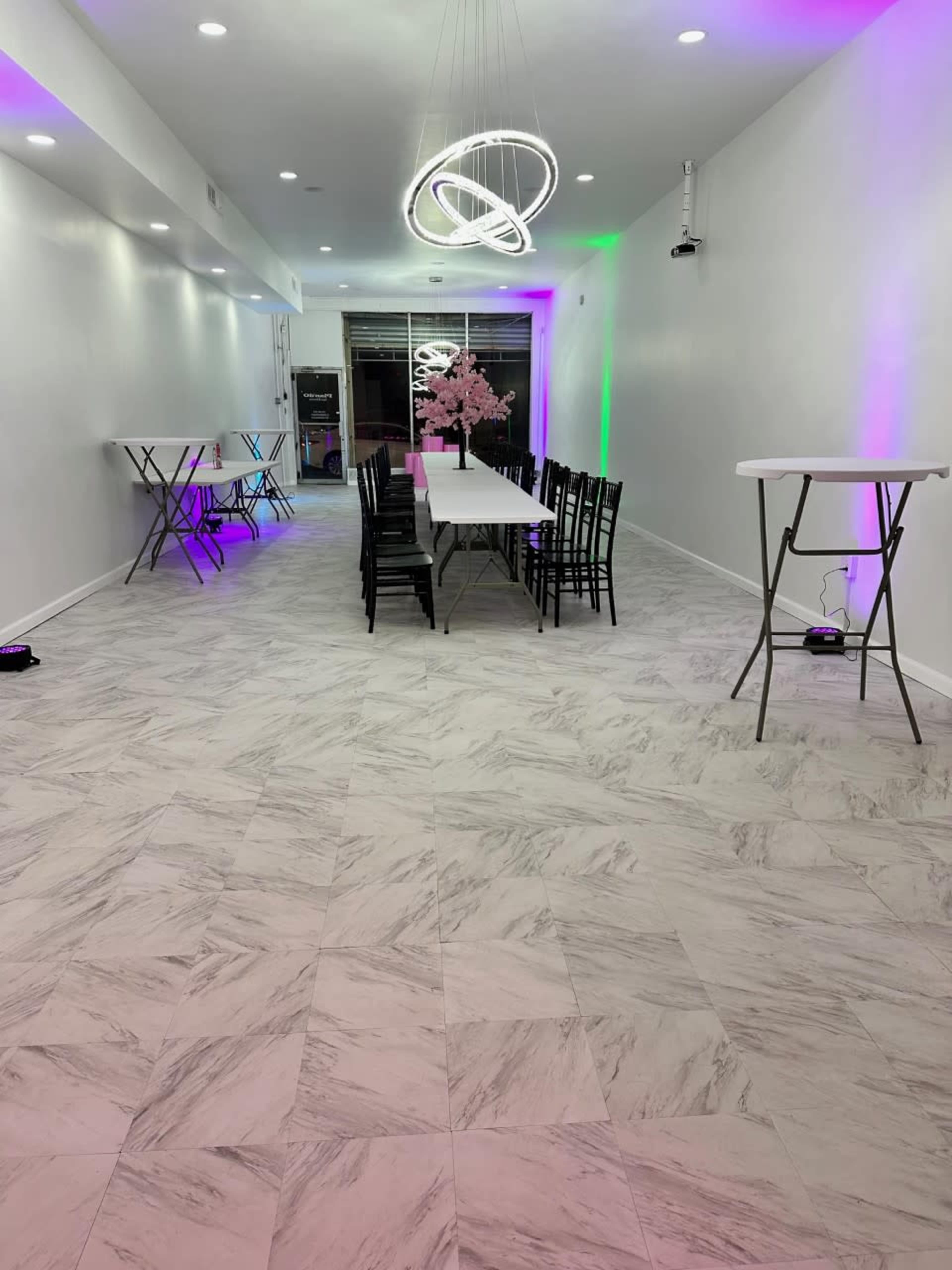 A spacious white room features a long dining table with black chairs, illuminated by a modern chandelier, and provides a view of a pink floral arrangement.
