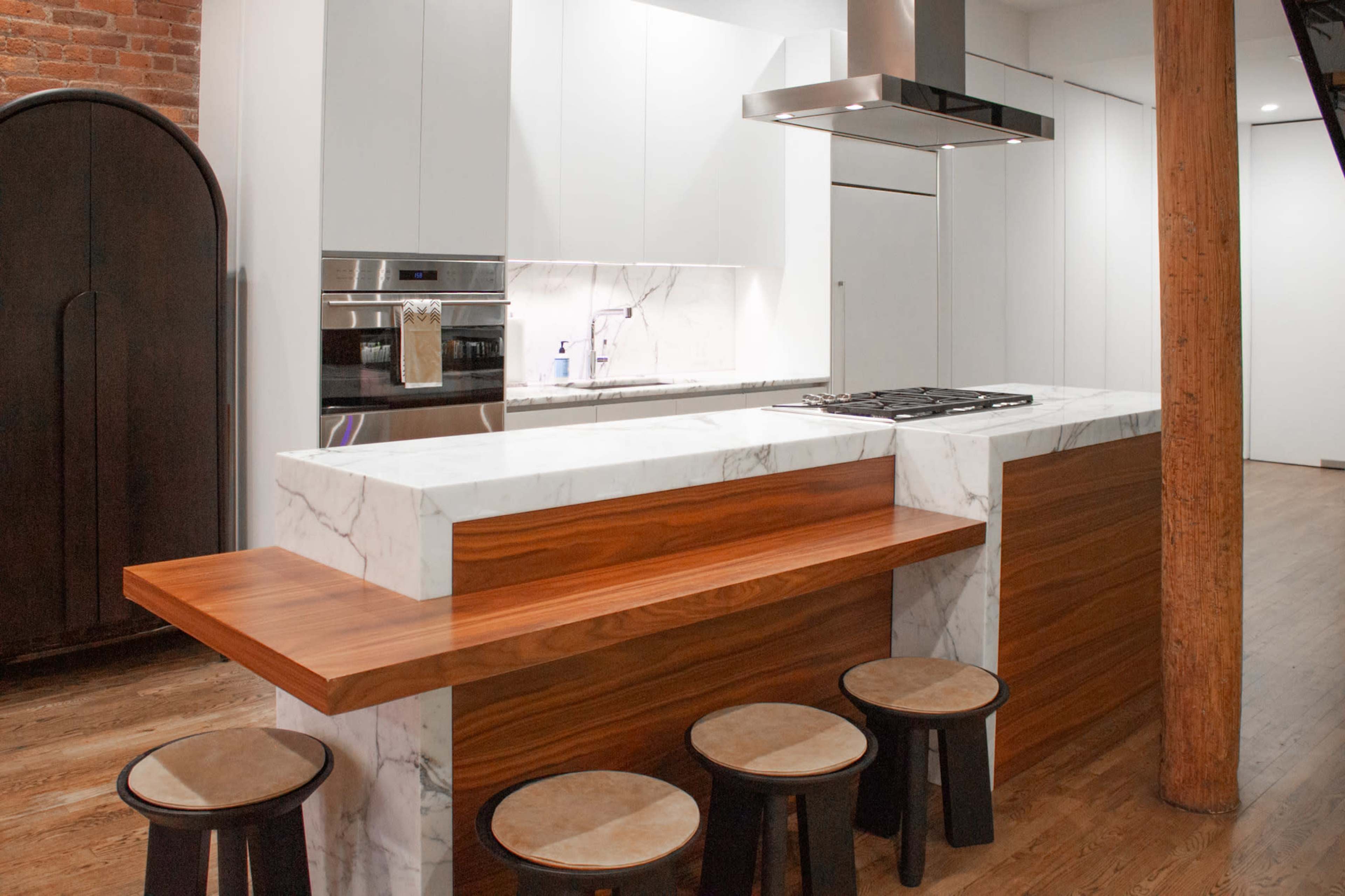 The image shows a modern kitchen featuring a marble-topped island with a wooden seating area and several stools, alongside sleek cabinetry and stainless steel appliances.