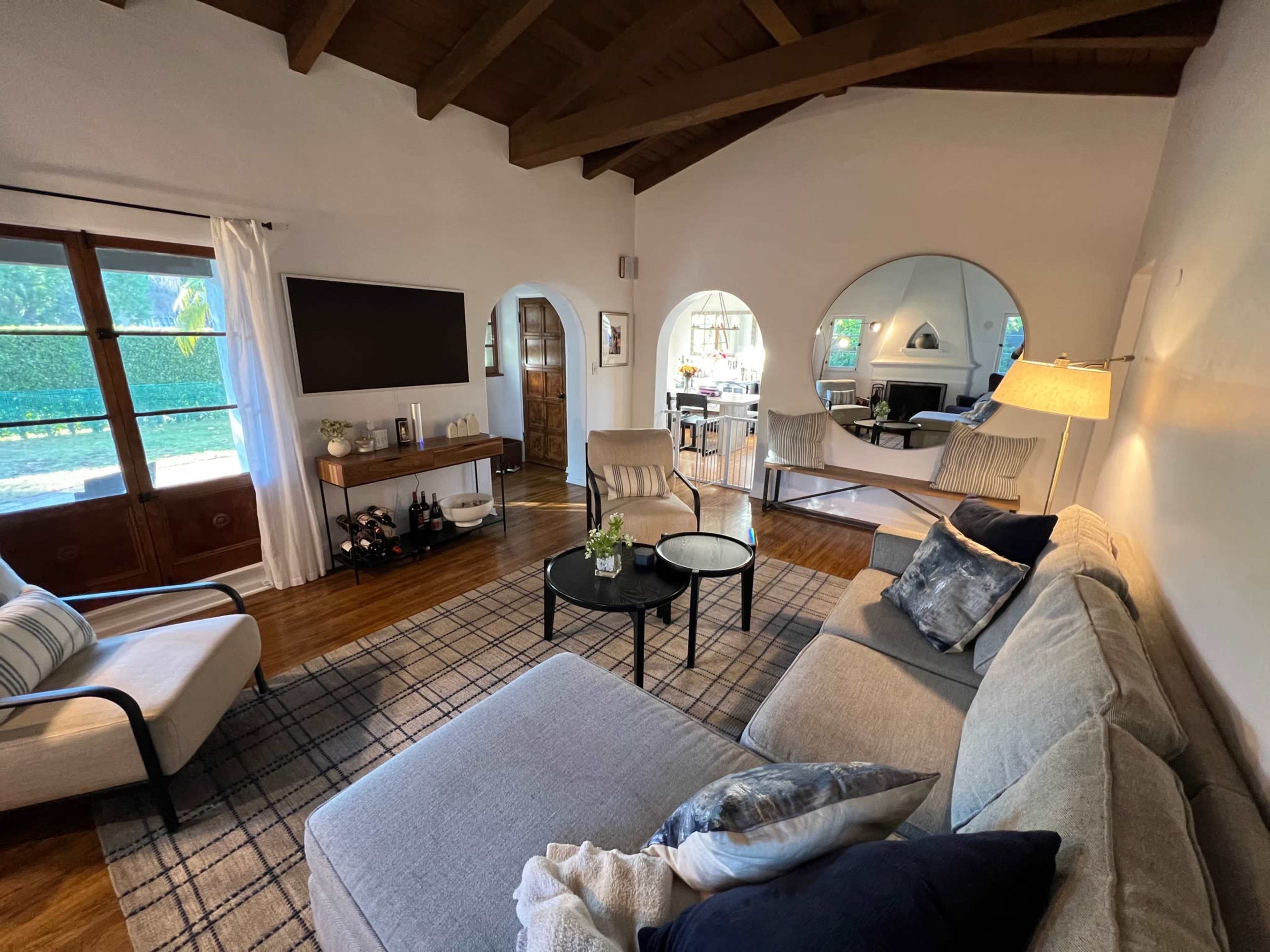 The image shows a cozy living room with large windows, a sectional sofa, a coffee table, and round mirrors on the wall.