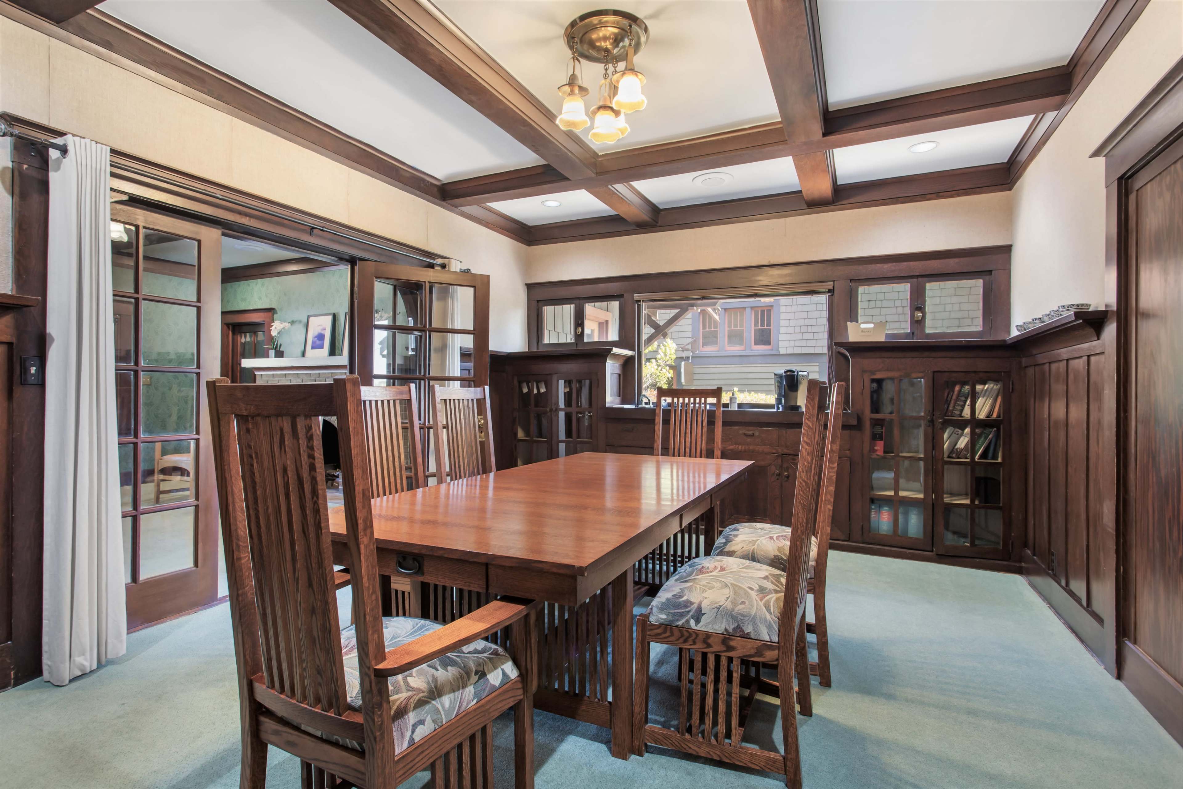 A wooden dining room with a large table surrounded by six chairs, large windows allowing natural light, and built-in shelves against the walls.