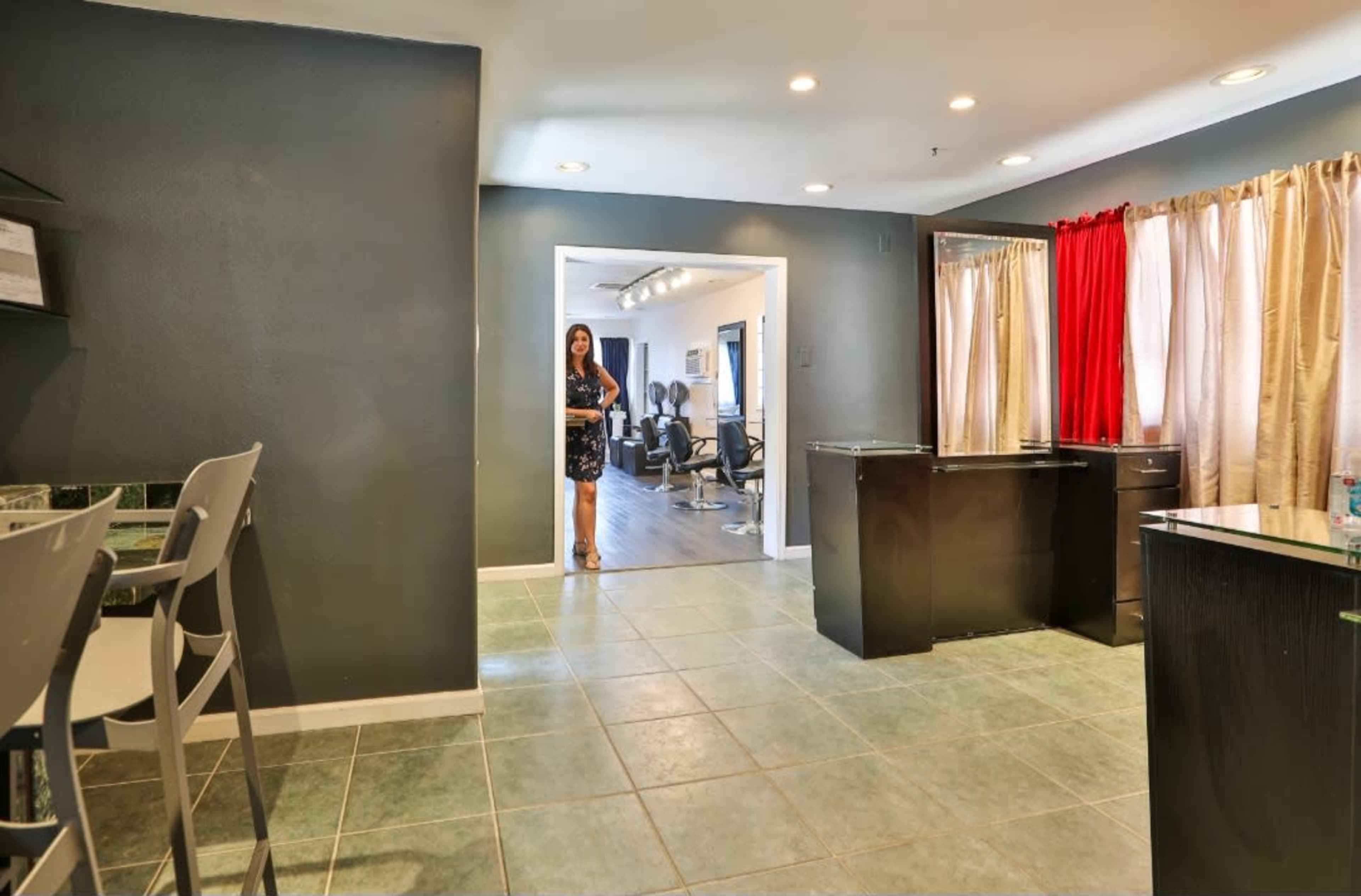 The image shows a spacious room with a kitchen area featuring a tiled floor, bar stools, and a woman standing in a doorway leading to another room with salon chairs.
