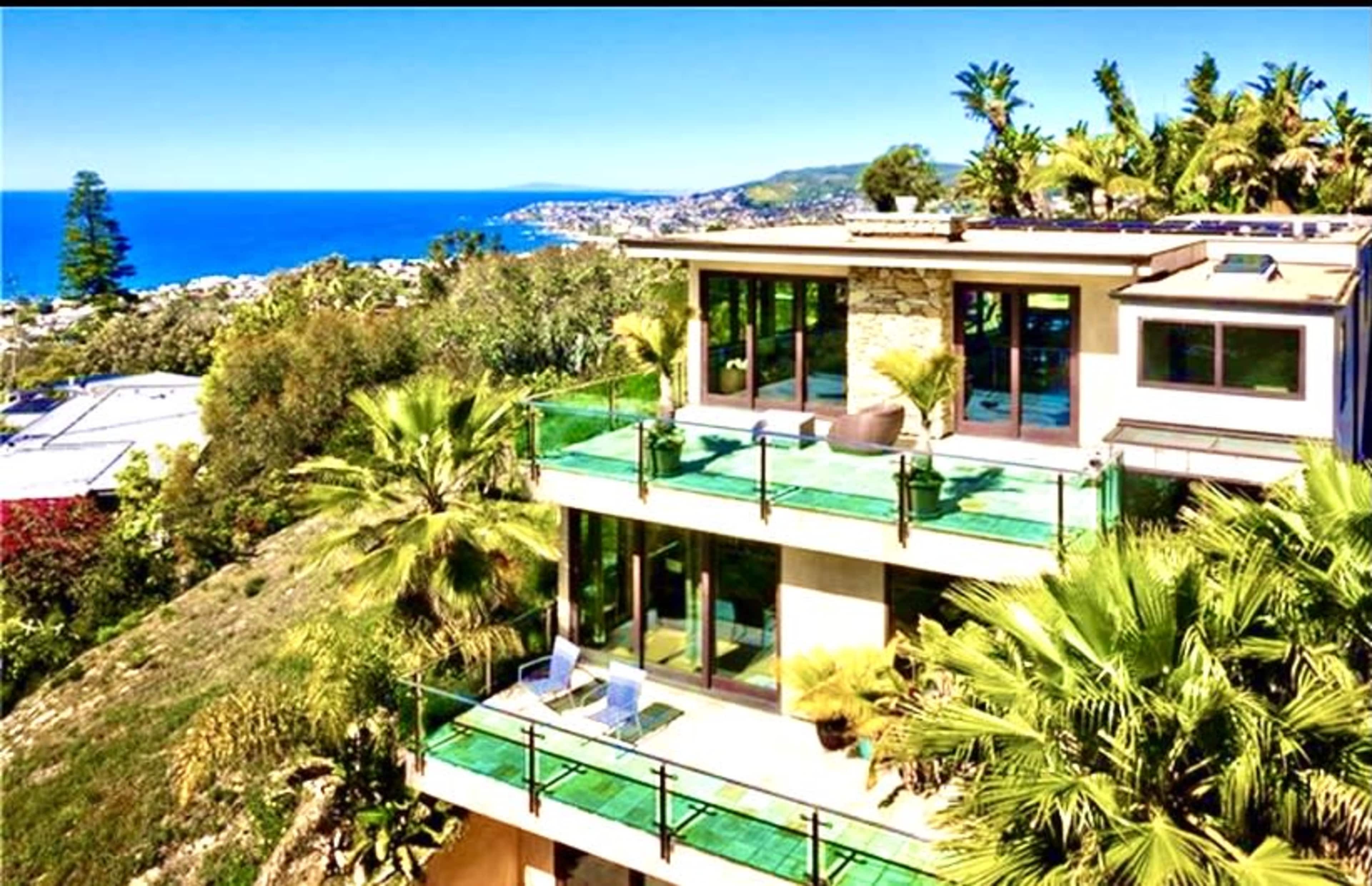 A modern house with glass balconies overlooks a coastal view and surrounding greenery.
