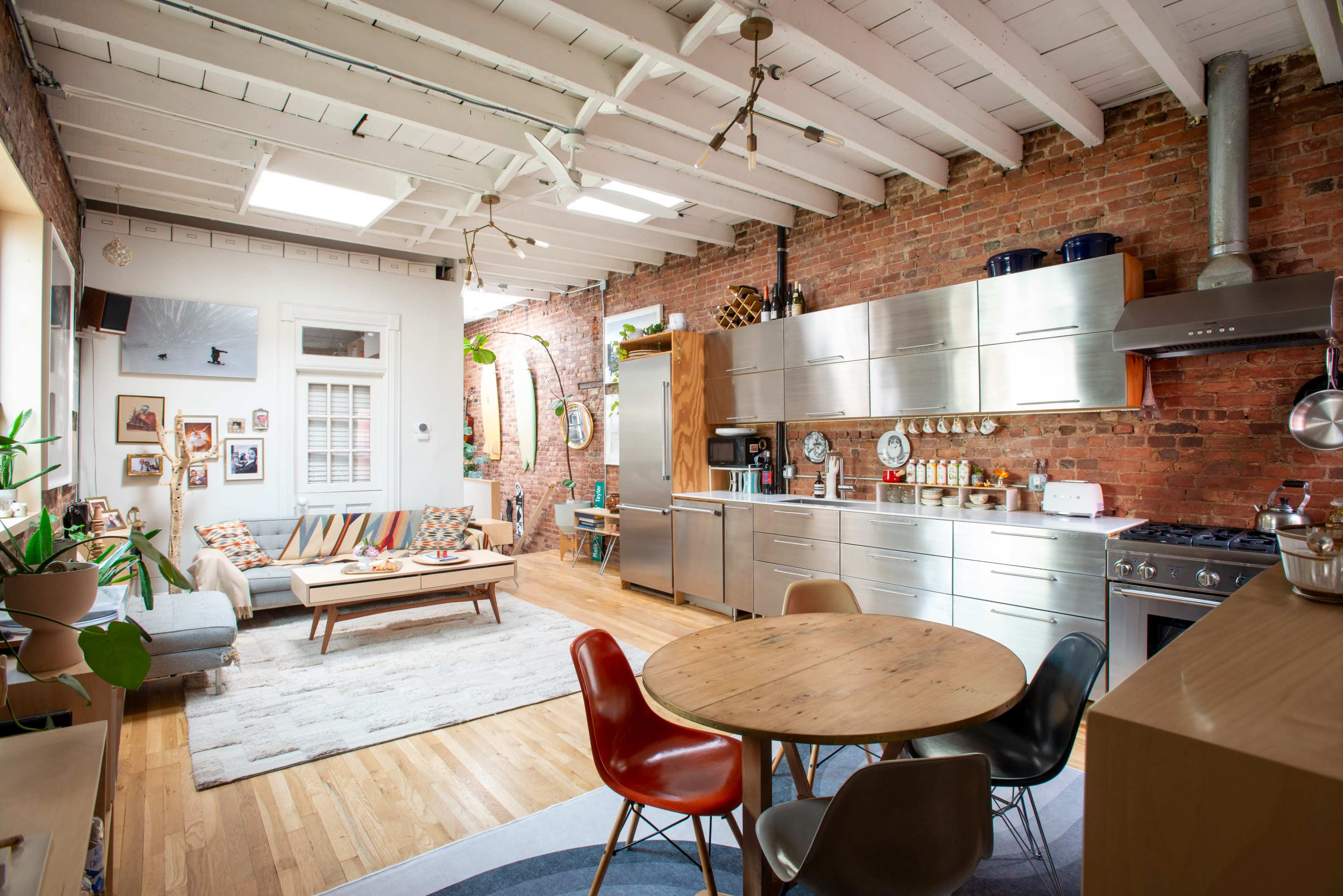 The image depicts a stylish kitchen and living area featuring exposed brick walls, modern stainless steel appliances, a wooden dining table with colorful chairs, and a cozy seating arrangement.