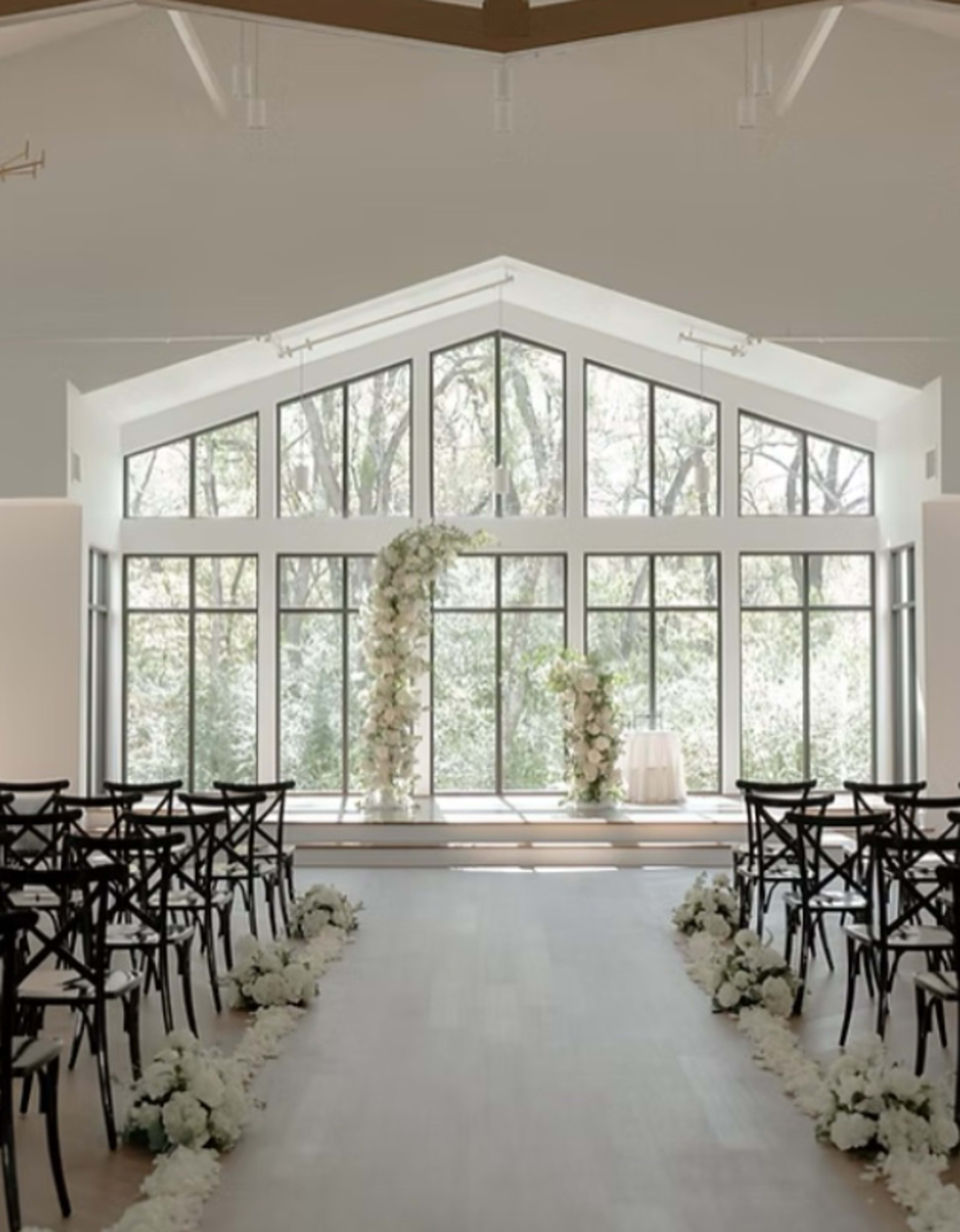 A modern wedding venue features a large row of windows overlooking a wooded area, with a floral arch at the front and chairs arranged on either side of a center aisle.