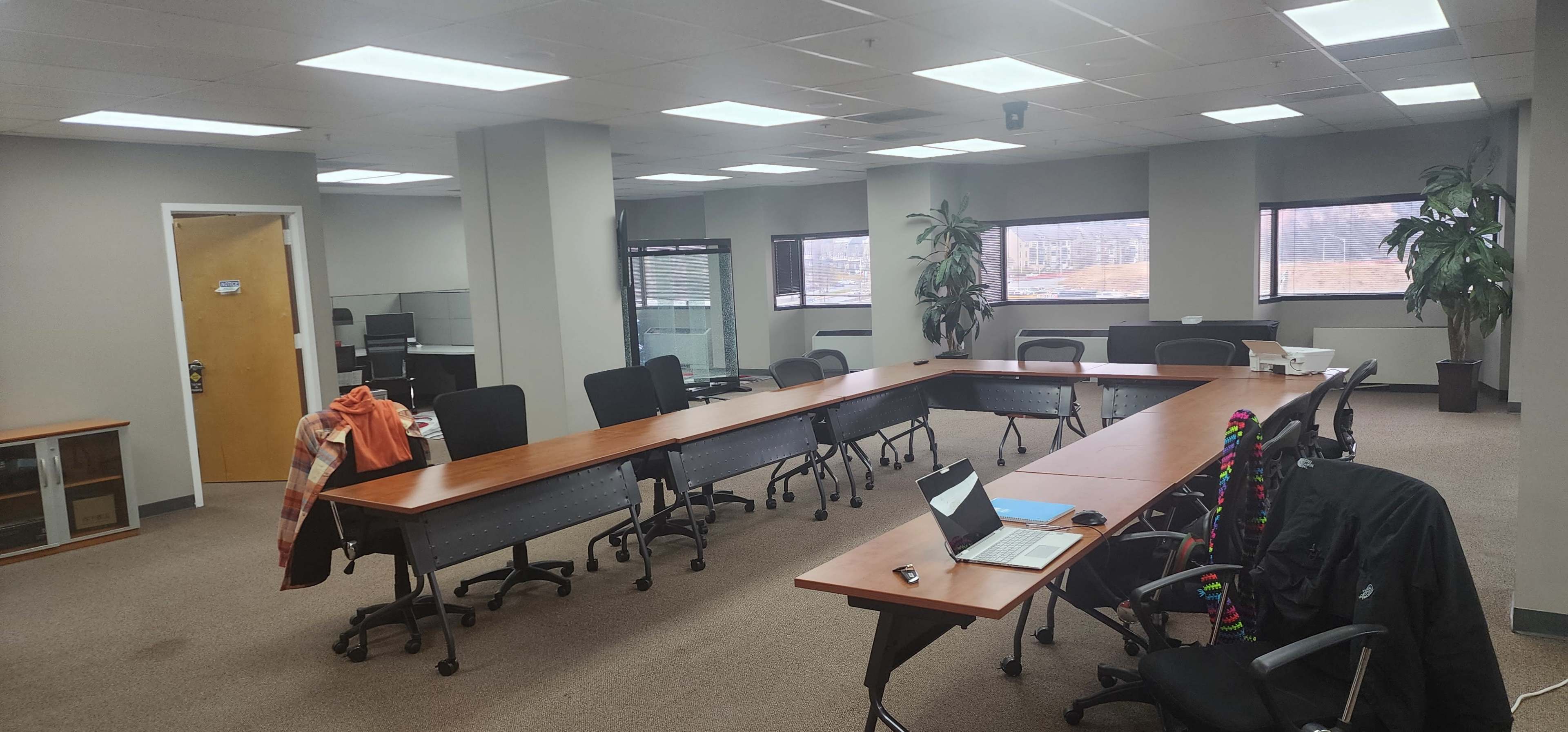 A spacious, modern office with a large conference table surrounded by chairs, a laptop on the table, and large windows revealing a view of the outdoors.