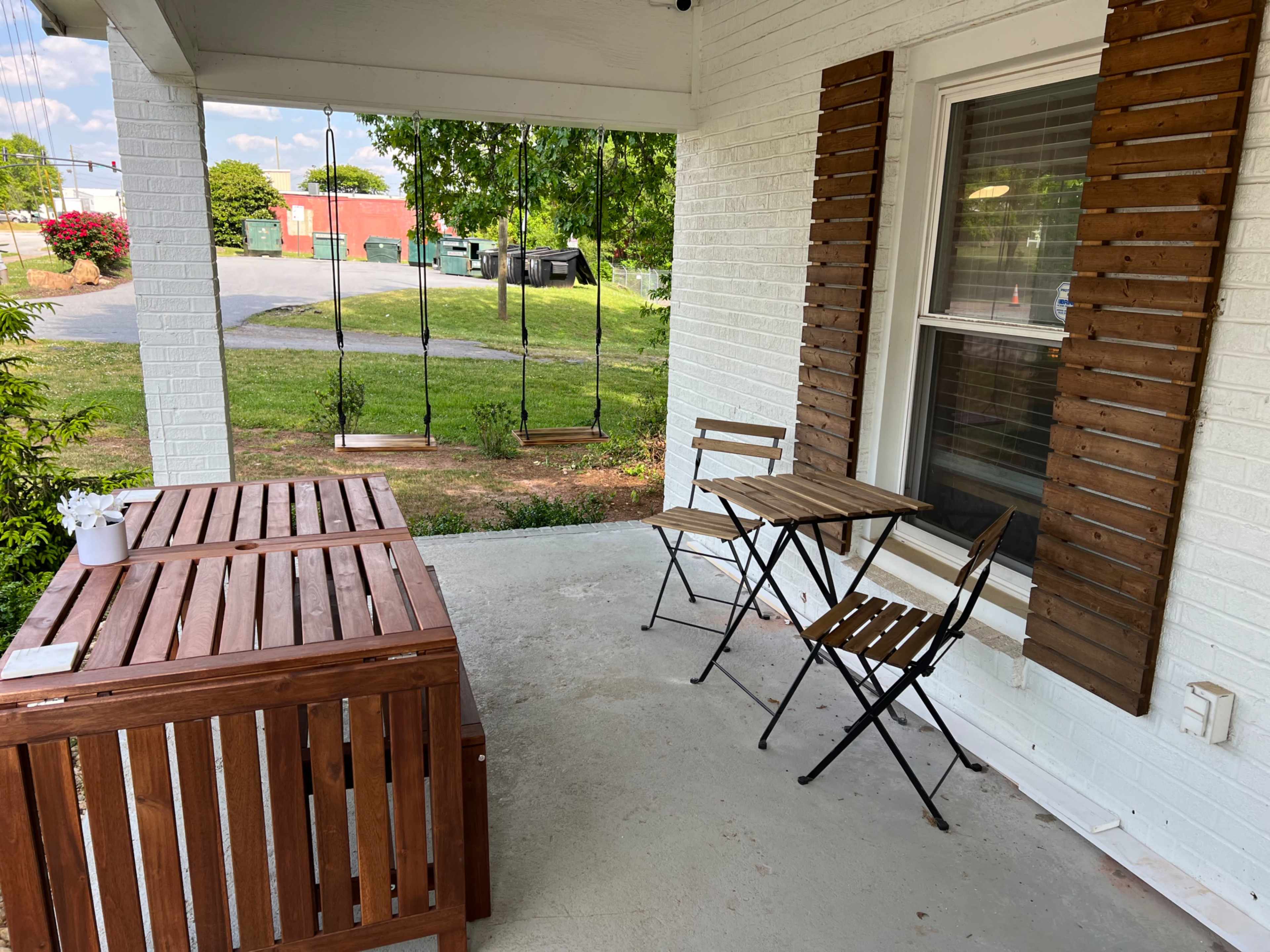The image shows a porch area with two folding chairs and a small table beside a large wooden storage bench, set against a backdrop of green grass and a distant parking area.
