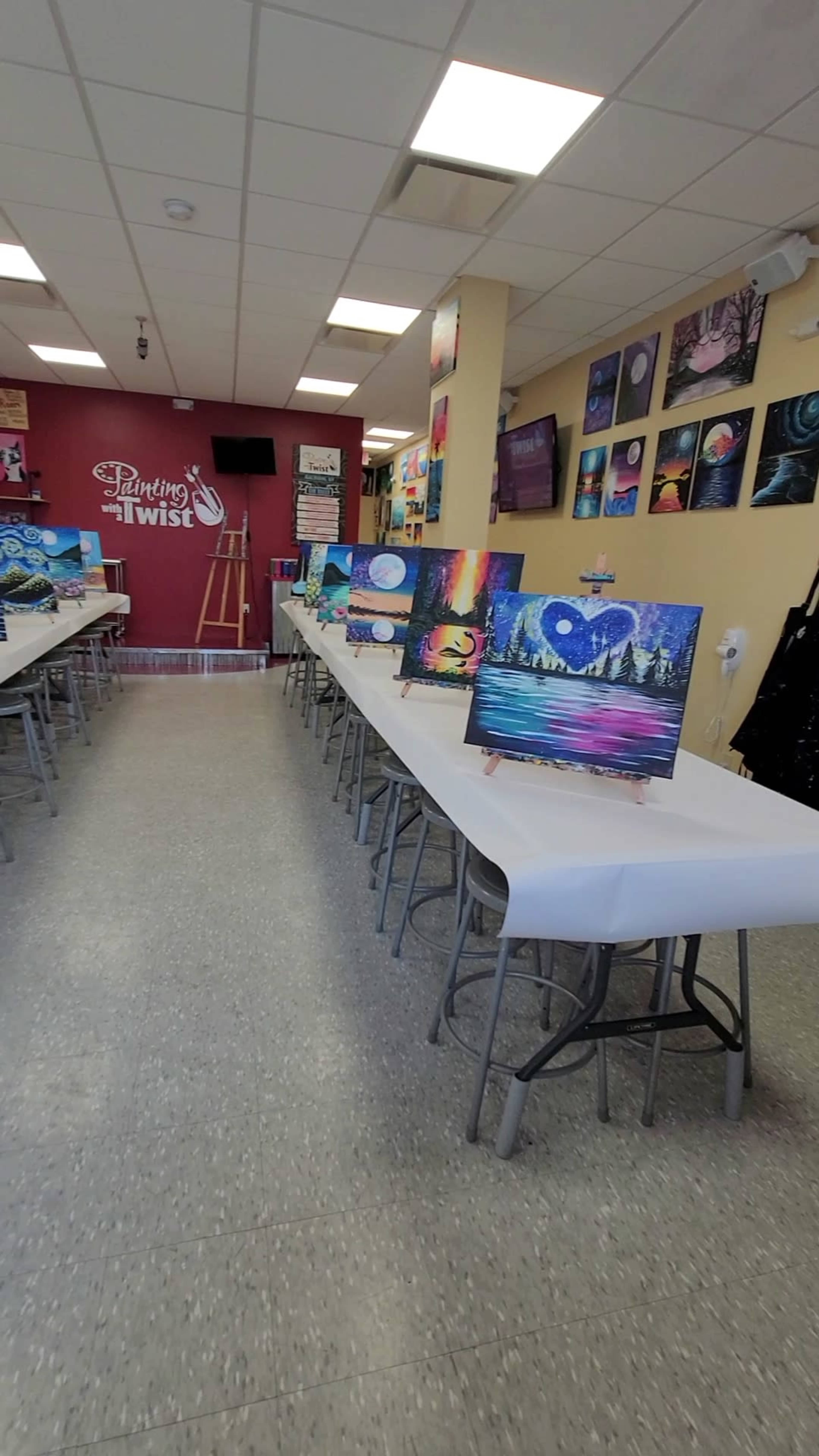 The image shows an art studio with several tables arranged in rows, each displaying a finished painting on an easel, and walls adorned with colorful artwork.