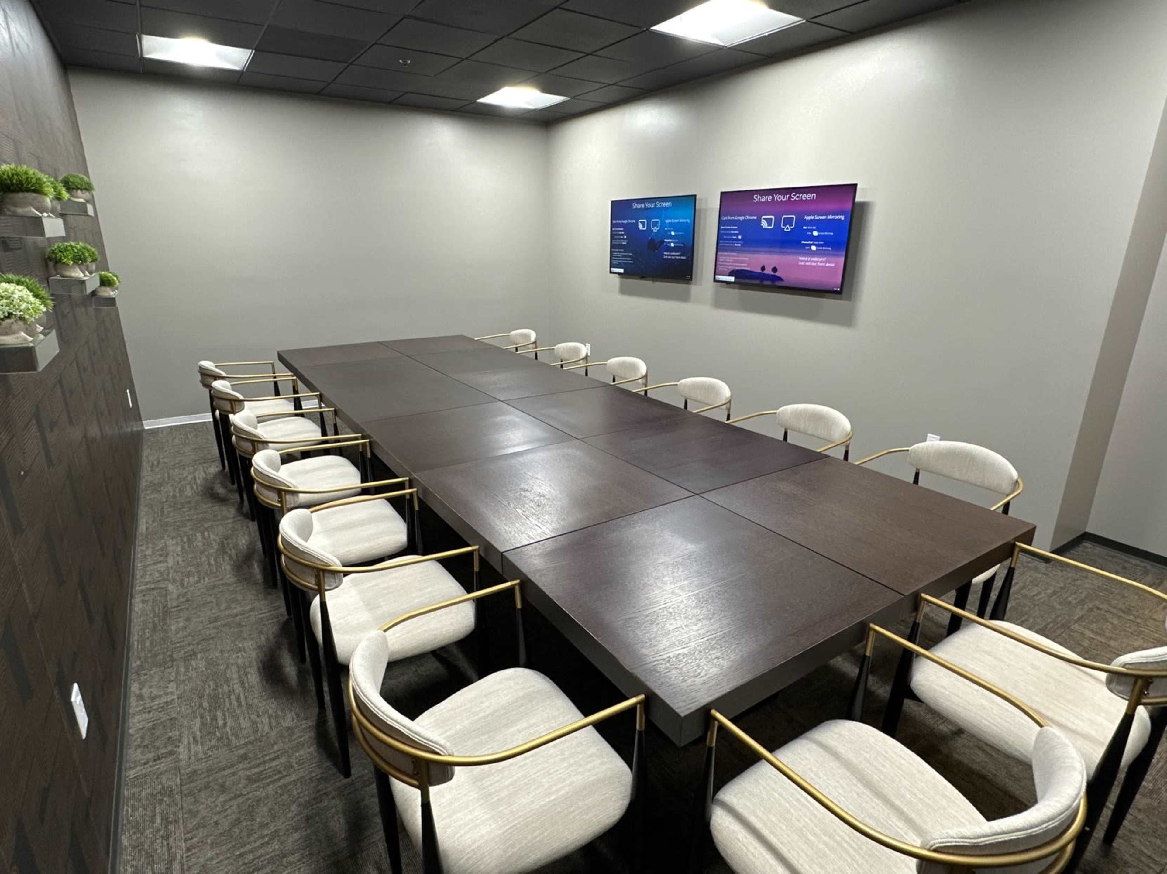The image shows a modern conference room with a long wooden table surrounded by twelve chairs and two screens mounted on the wall.