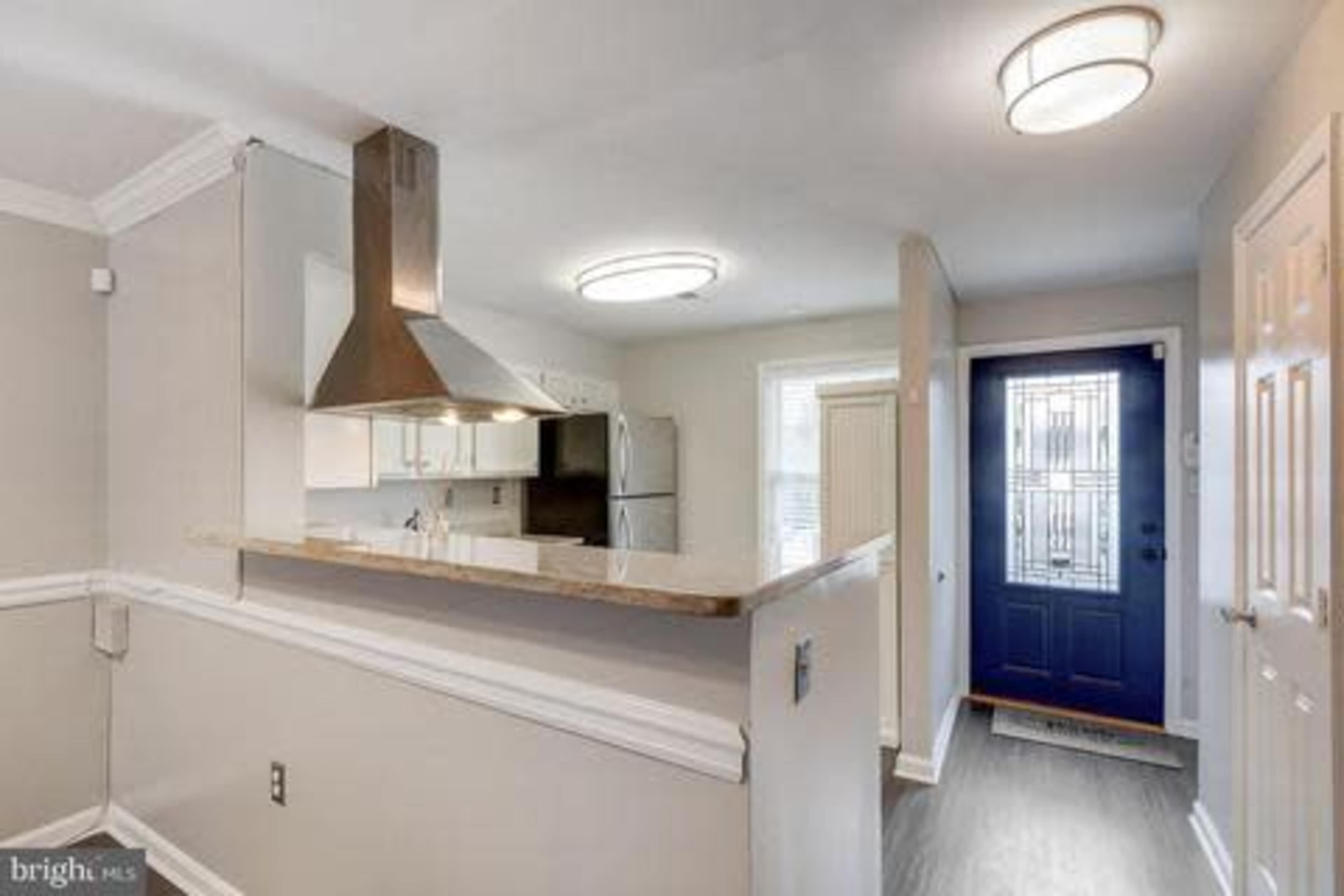The image shows a modern kitchen area with a stainless steel range hood, white cabinets, and a blue front door leading to an entryway.