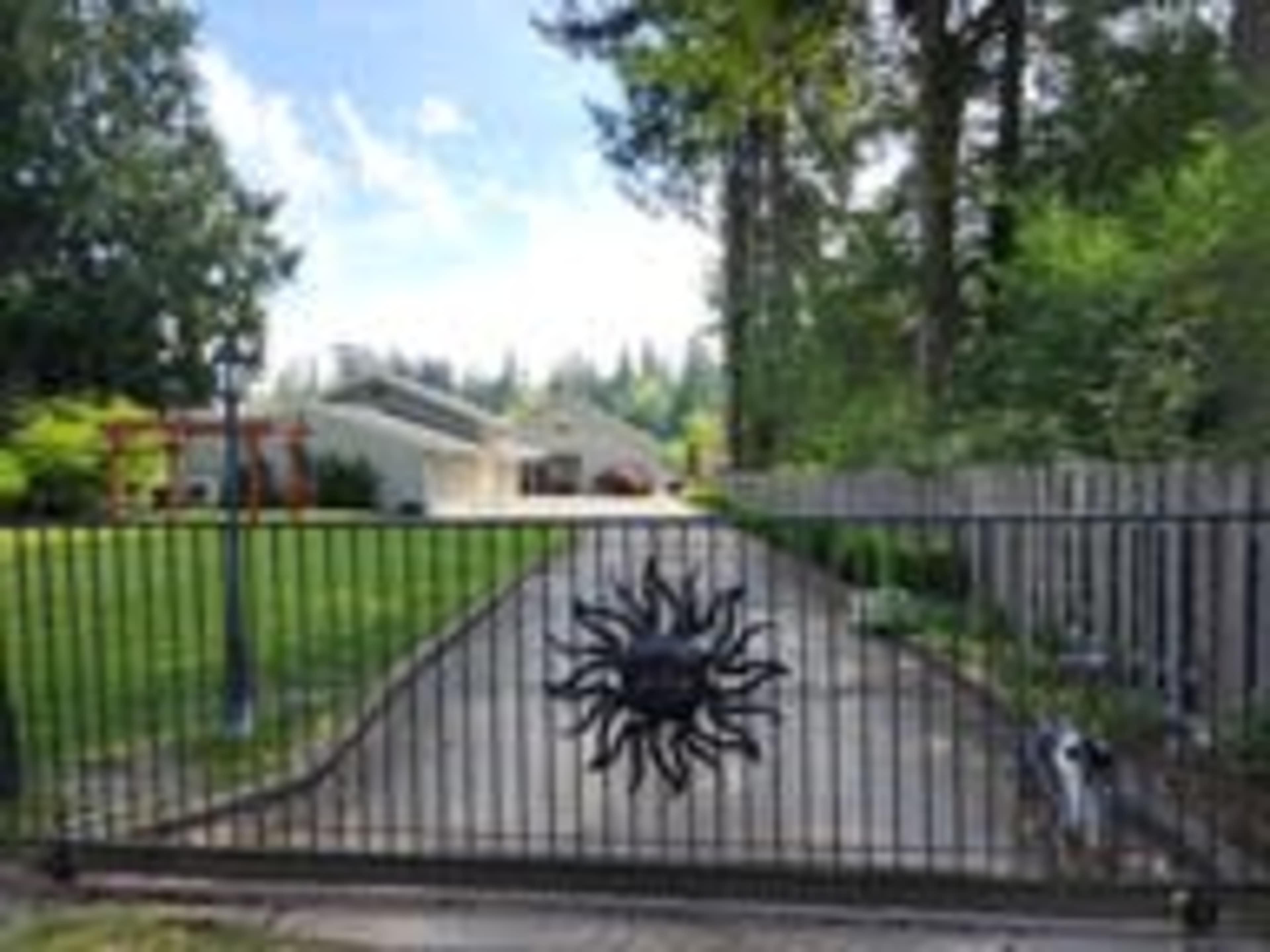 A wrought iron gate featuring a sun design opens to a driveway leading to a house surrounded by trees.