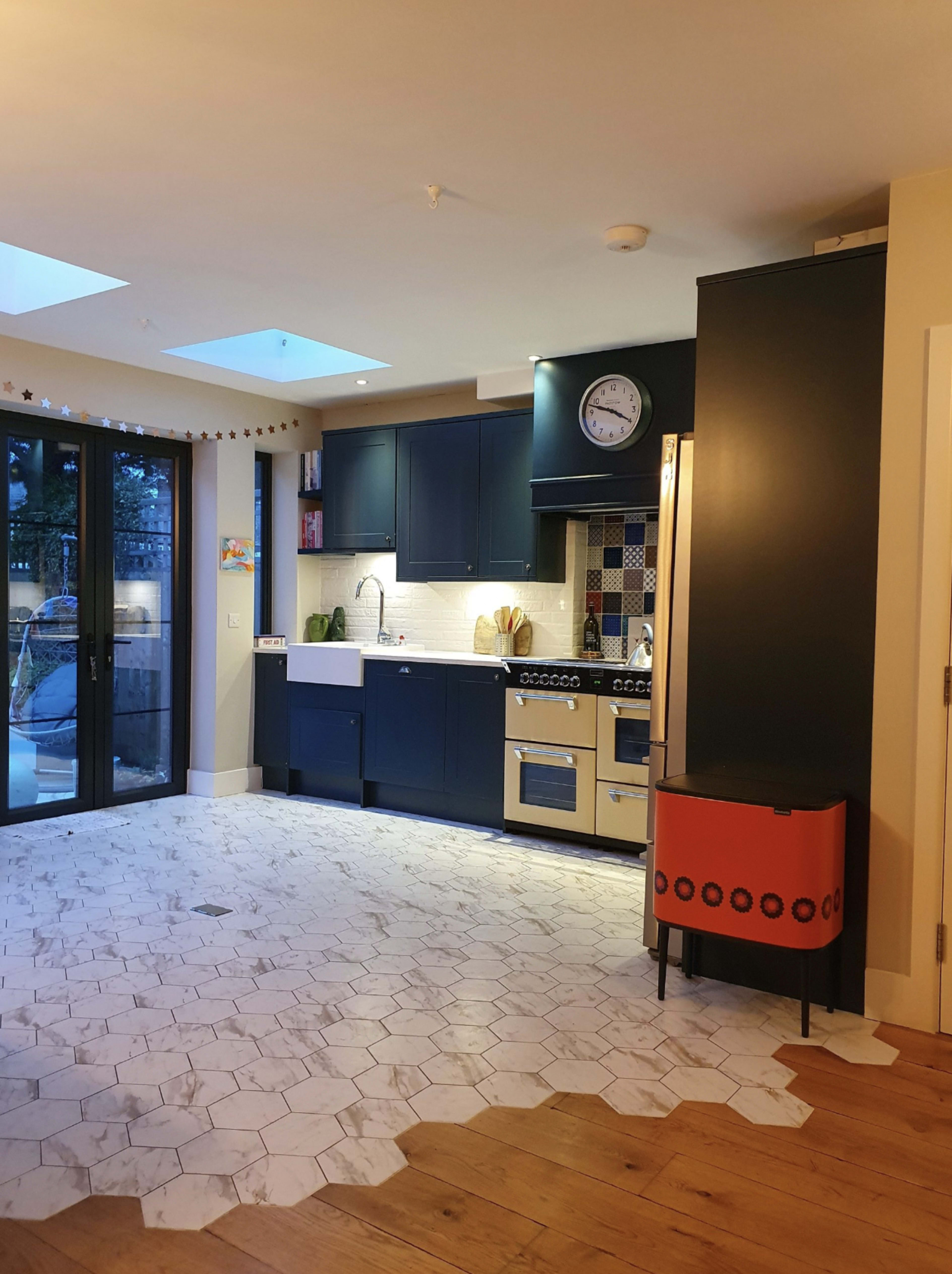The image shows a modern kitchen with blue cabinets, a white farmhouse sink, and a mix of hexagonal and wooden flooring, featuring large glass doors that lead to an outdoor area.
