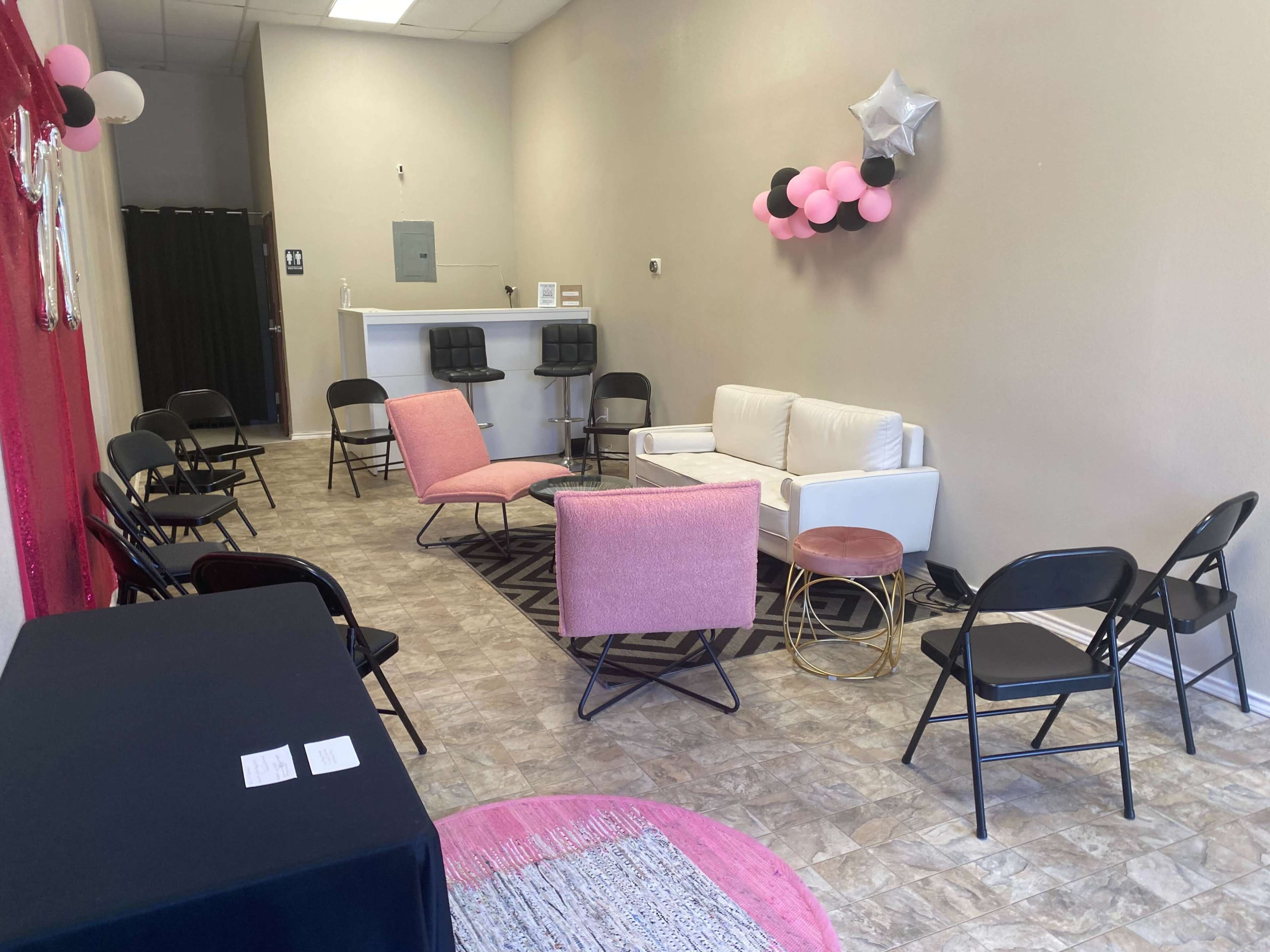 A small event space featuring a few pink chairs, a white couch, a black table, and decorative balloons on the wall.