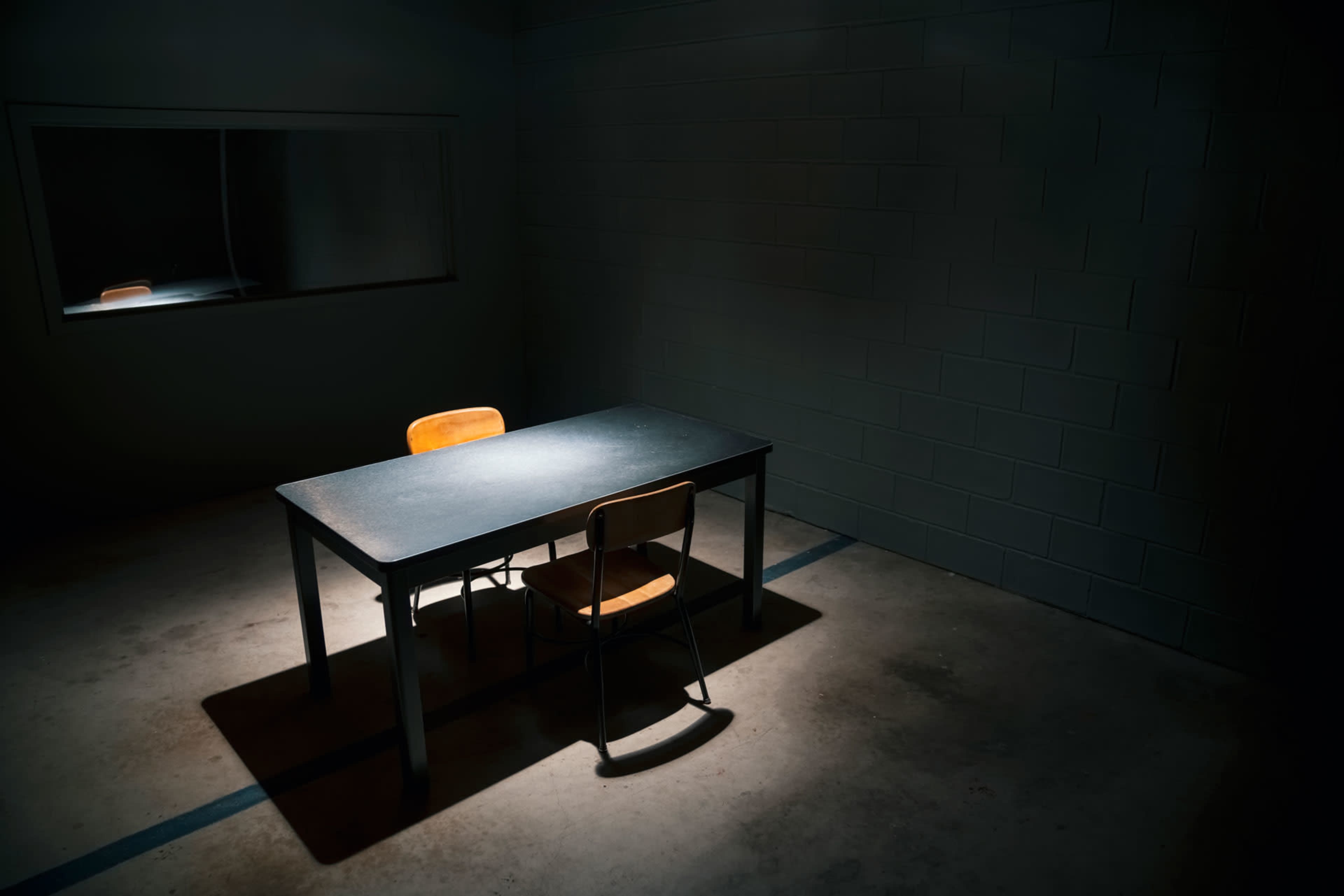 A dimly lit interrogation room features a lone table with two chairs positioned under a spotlight, while a small window reflects the faint light.