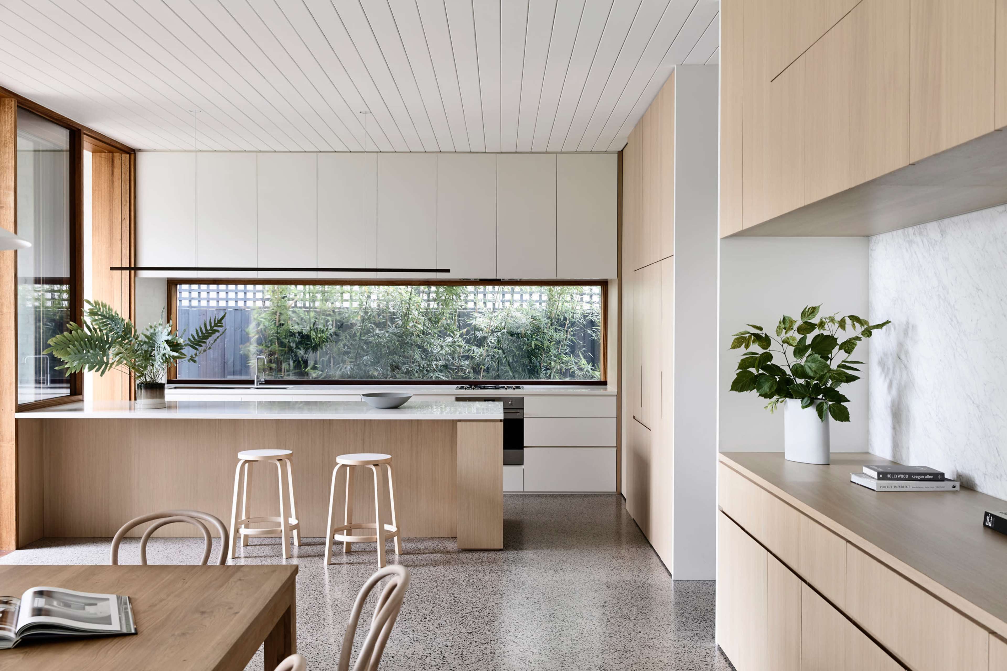 A modern kitchen with light wood cabinetry, a central island with three stools, and large windows overlooking a garden.