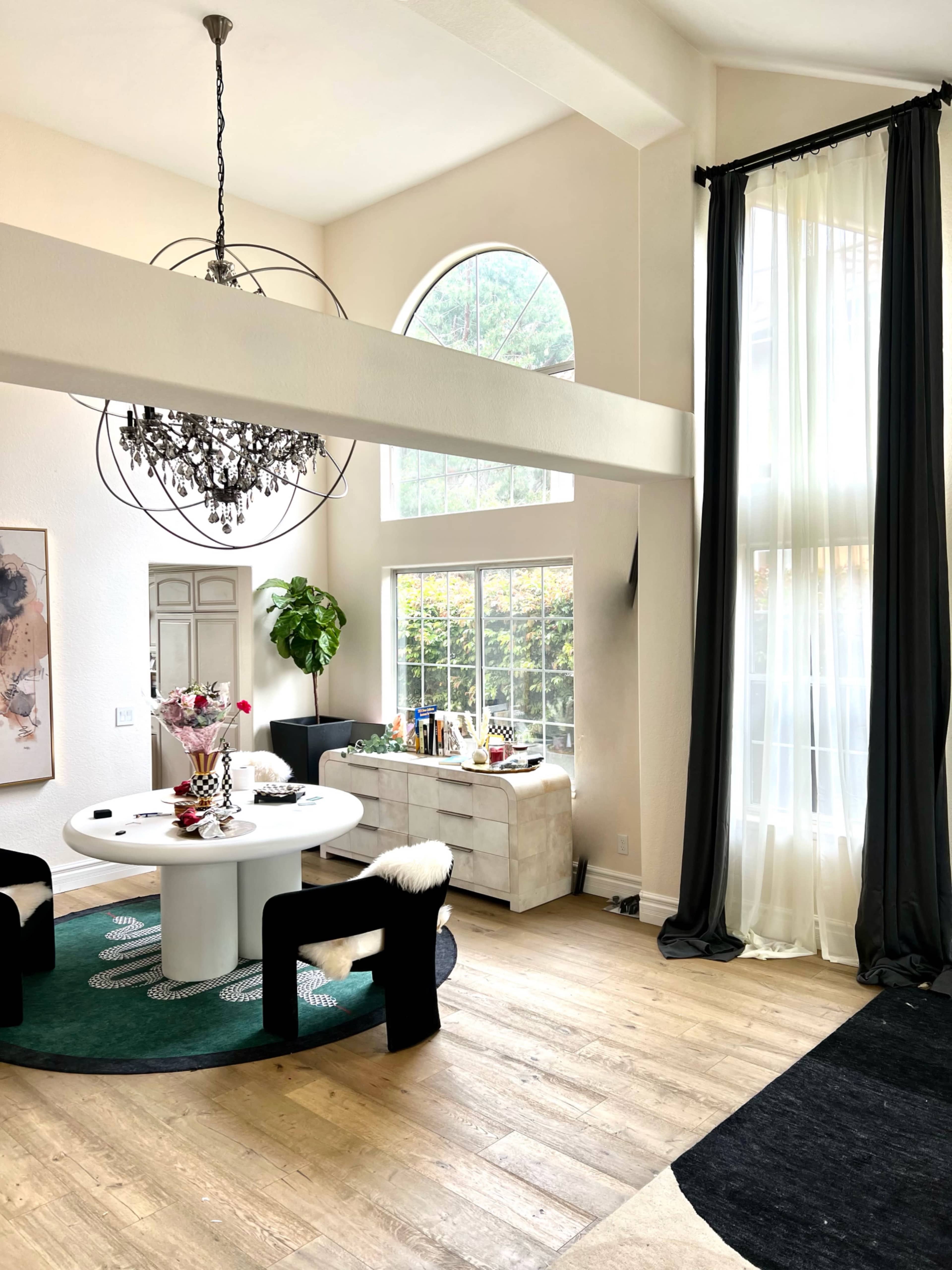 A modern dining area featuring a round table surrounded by black chairs, a large window with sheer curtains, and a chandelier hanging from the ceiling.