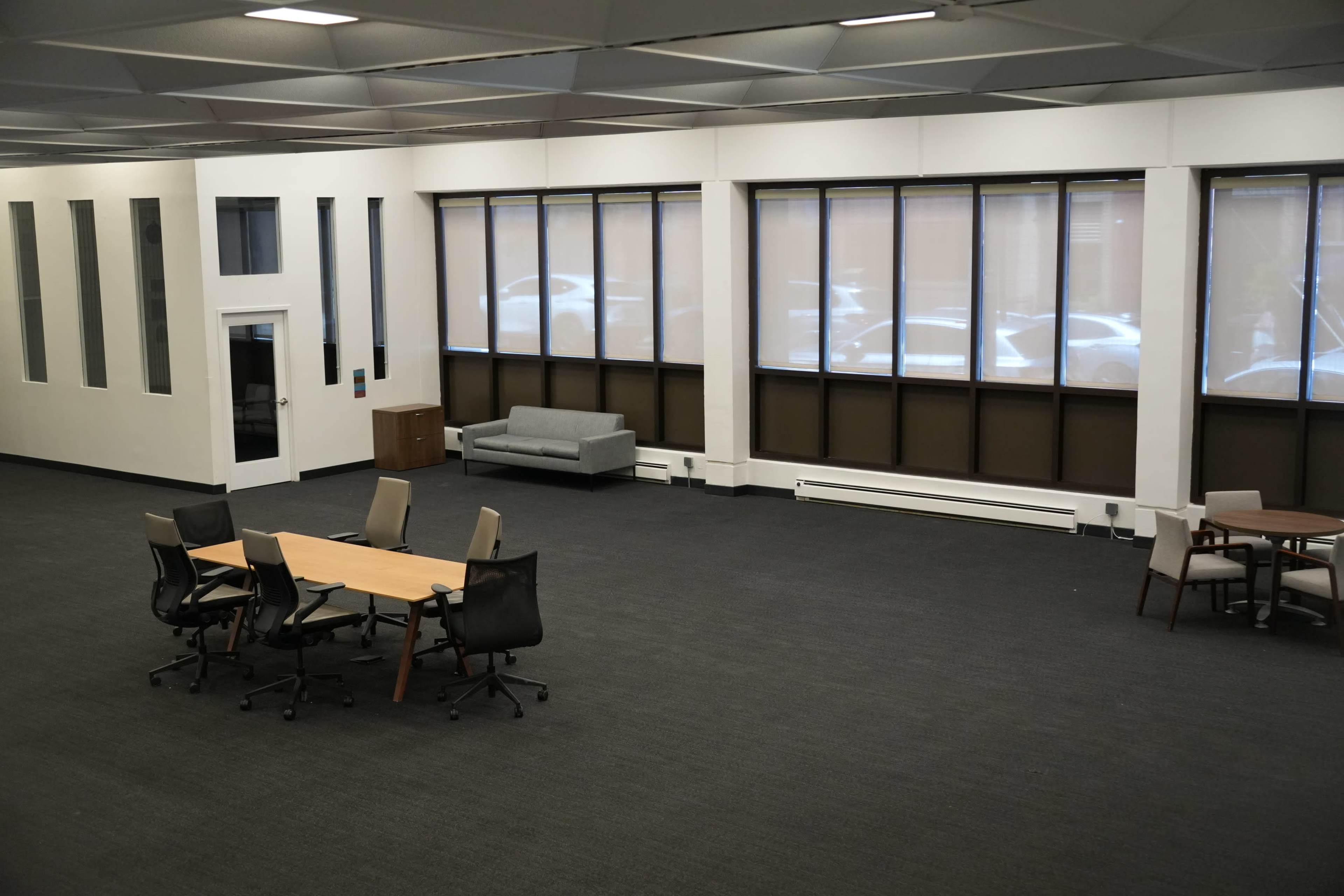 The image shows a spacious, empty room with large windows, a seating area with a gray couch, and a table surrounded by chairs.