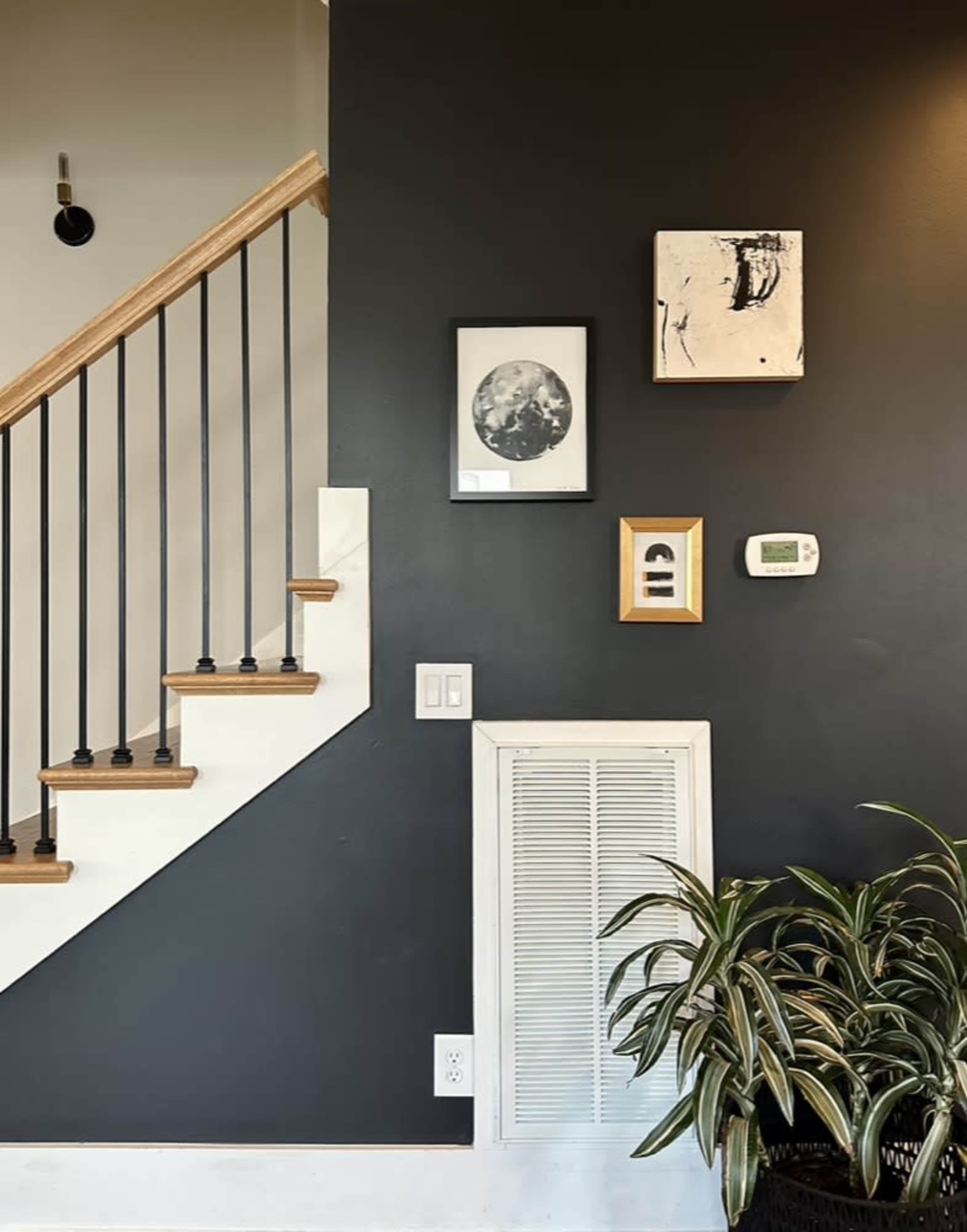 The image depicts a dark wall featuring framed art, a thermostat, and a potted plant beside a staircase.