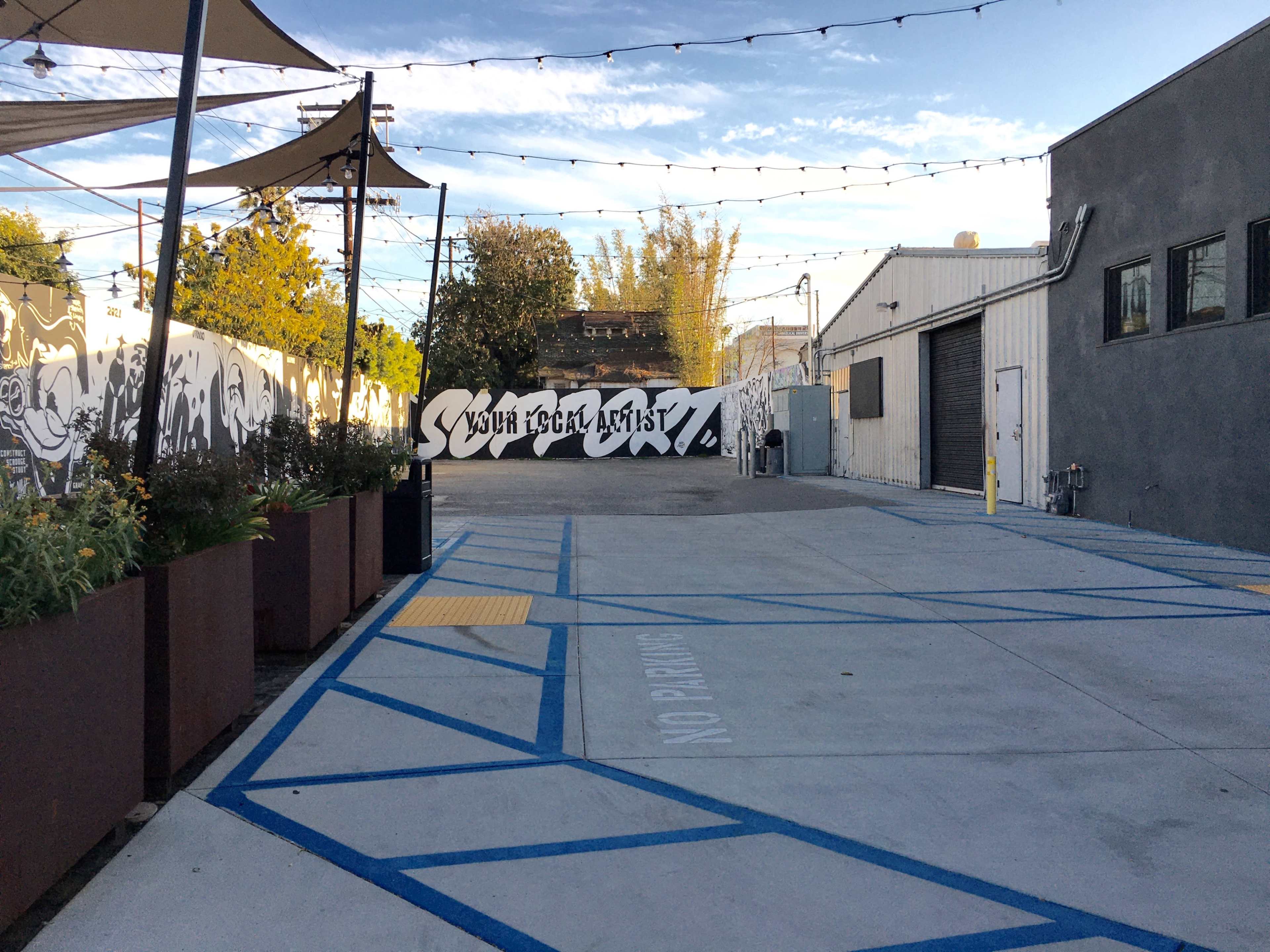 A paved outdoor area with a mural on the wall, surrounded by buildings and plants, under string lights.