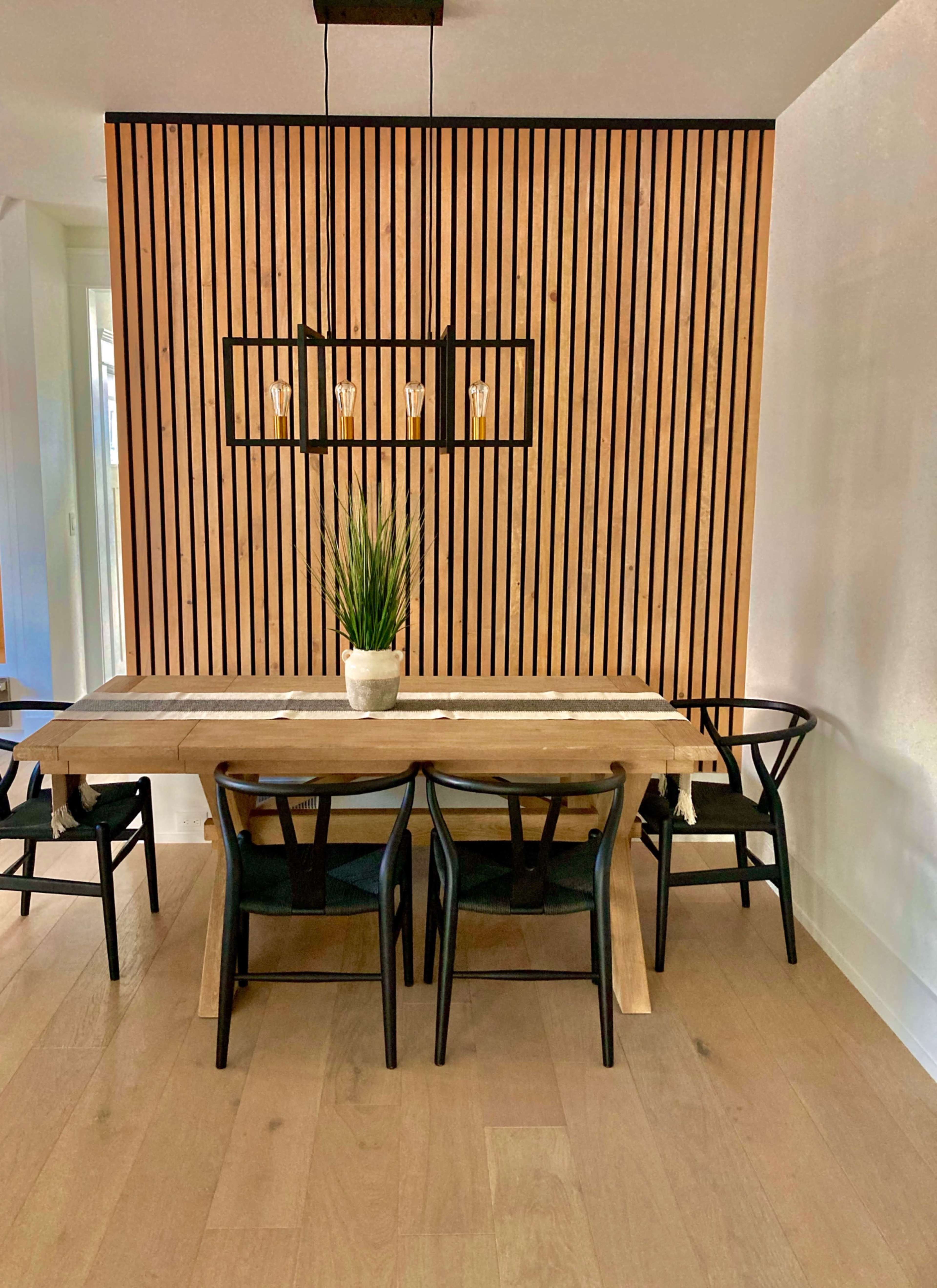 A wooden dining table with black chairs is set against a vertical slatted wood wall, illuminated by a rectangular light fixture above.