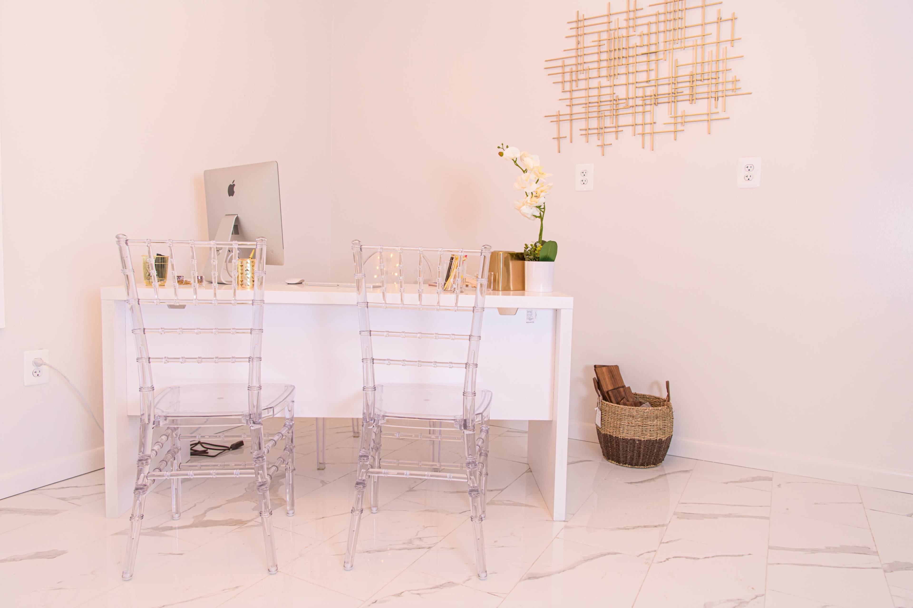 A minimalist home office features a white desk, two clear acrylic chairs, an iMac, and decorative items on a light-colored tiled floor.