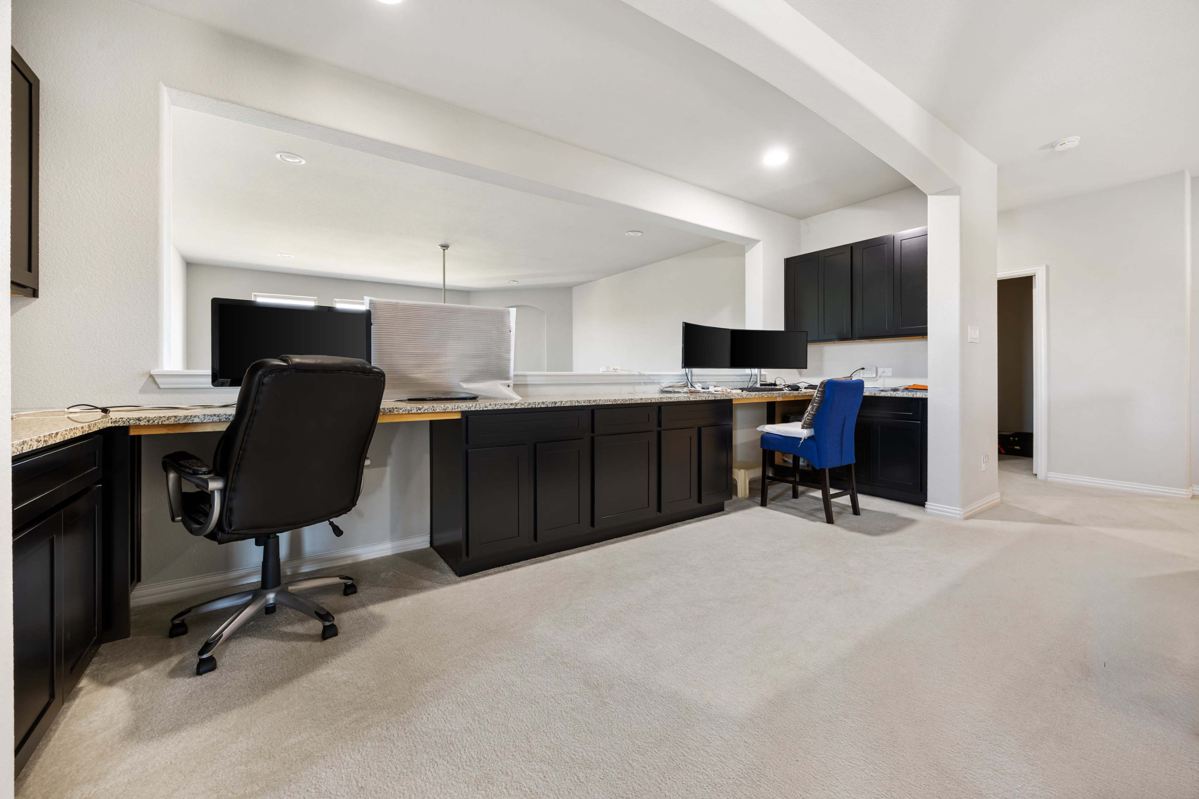 The image depicts a modern home office area featuring dark cabinetry, a granite countertop, two computer monitors, a black office chair, and a blue upholstered chair, all set on a beige carpet.