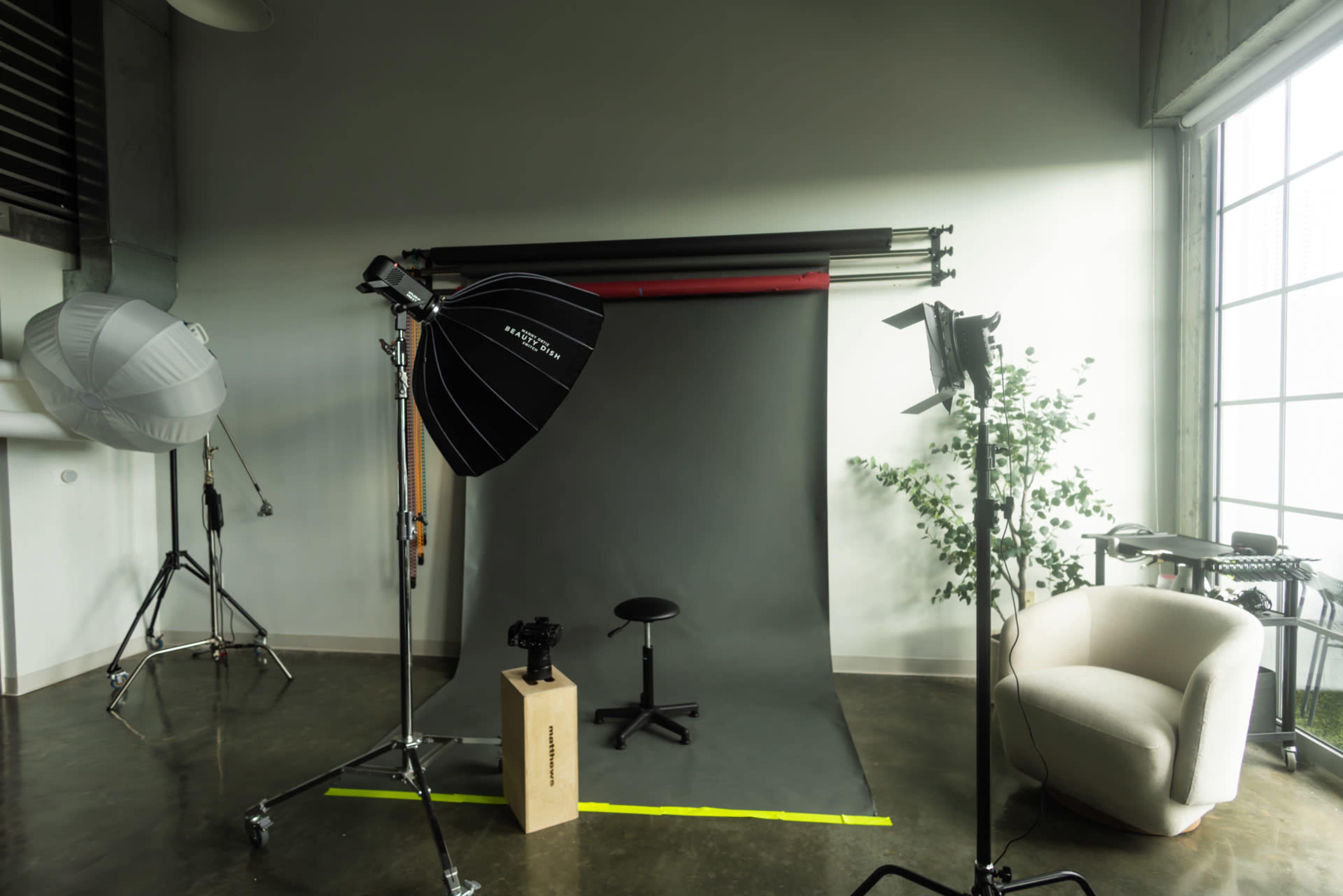 The image shows a photography studio with a gray backdrop, lighting equipment, a stool, and a chair positioned in a spacious, well-lit area.