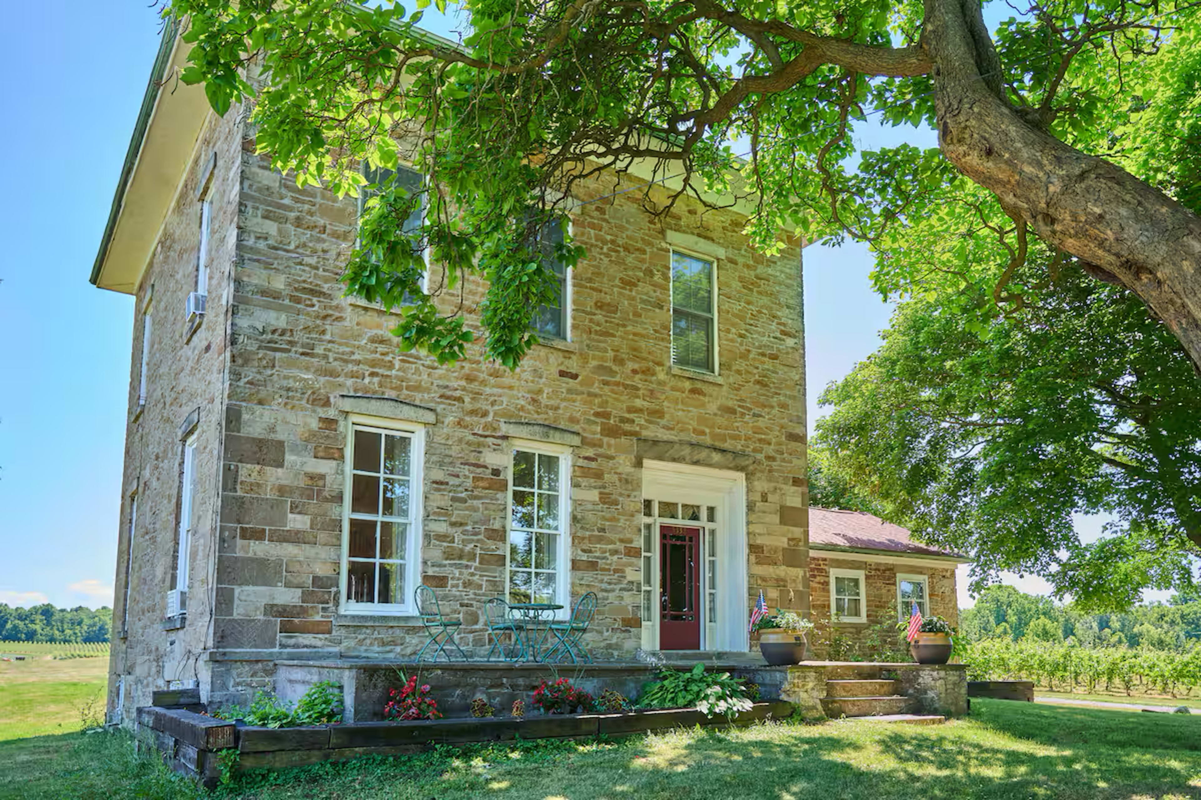 A two-story stone house with large windows and a front porch is surrounded by green trees and lawn, with a nearby smaller building visible in the background.