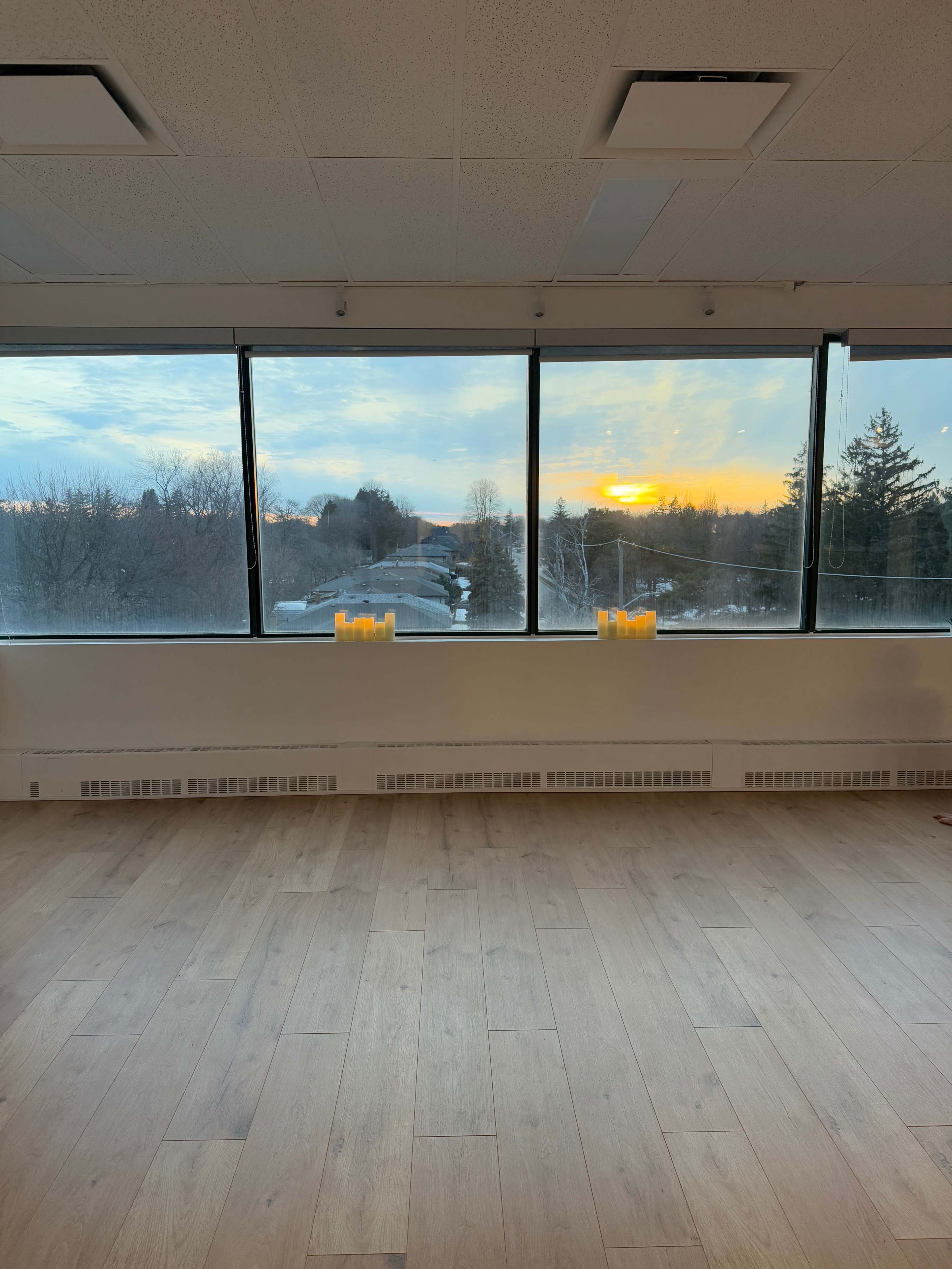 The image shows a spacious room with a wooden floor and large windows overlooking a sunset, with candles arranged on the floor in front of the windows.