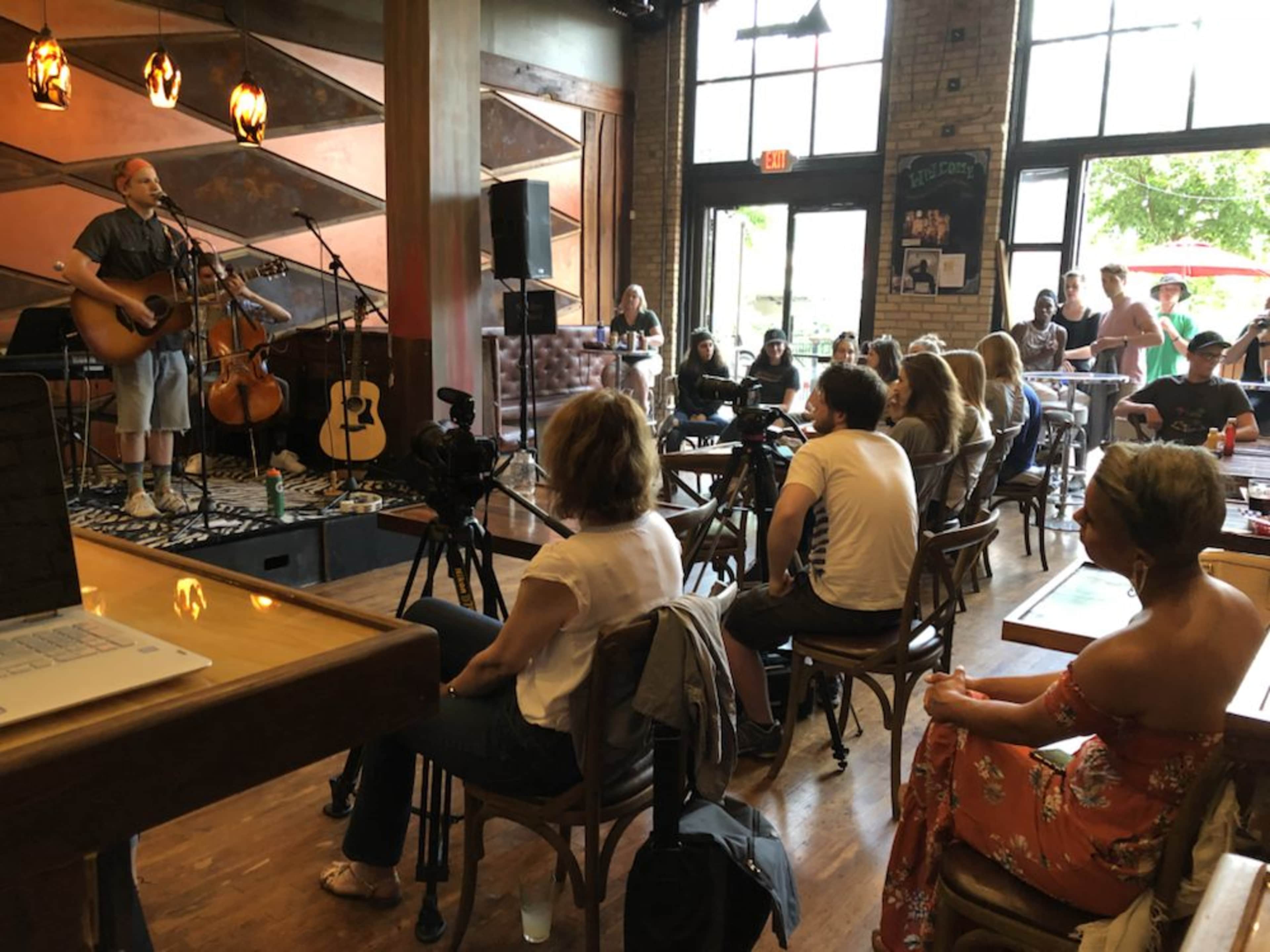A musician performs on stage with a guitar and a cello in a crowded café filled with seated audience members and camera equipment.