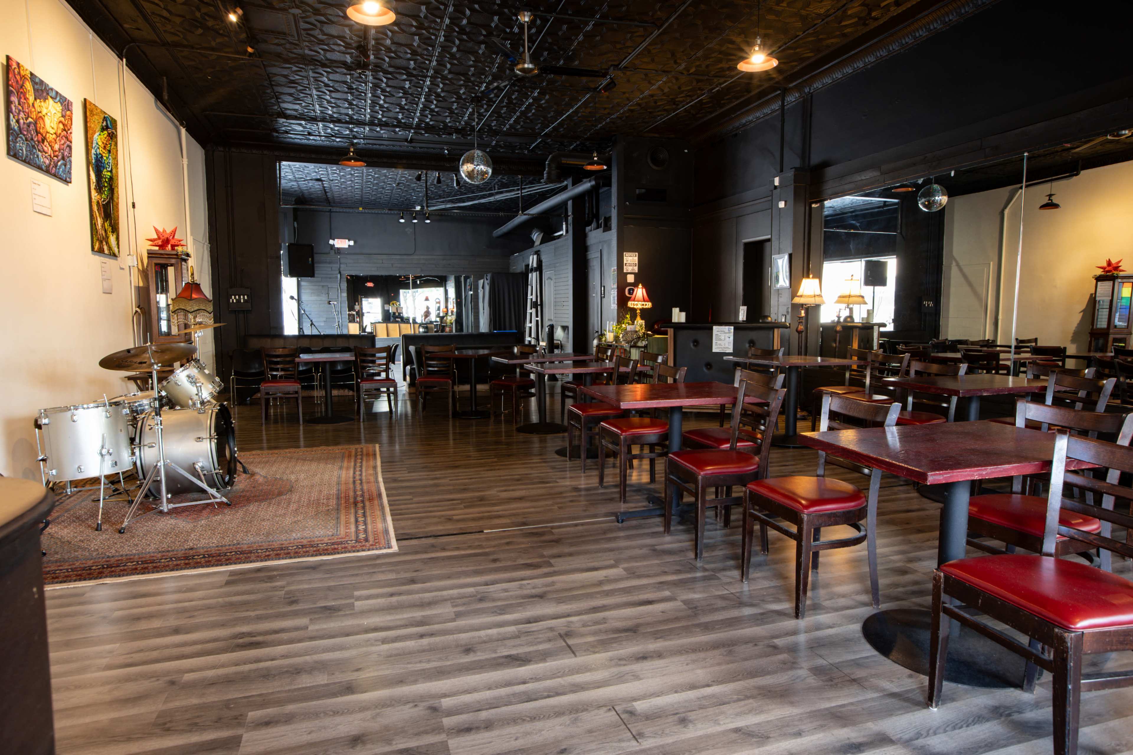 The interior of a restaurant features wooden tables and chairs, a small stage with a drum set, and decorative artwork on the walls.