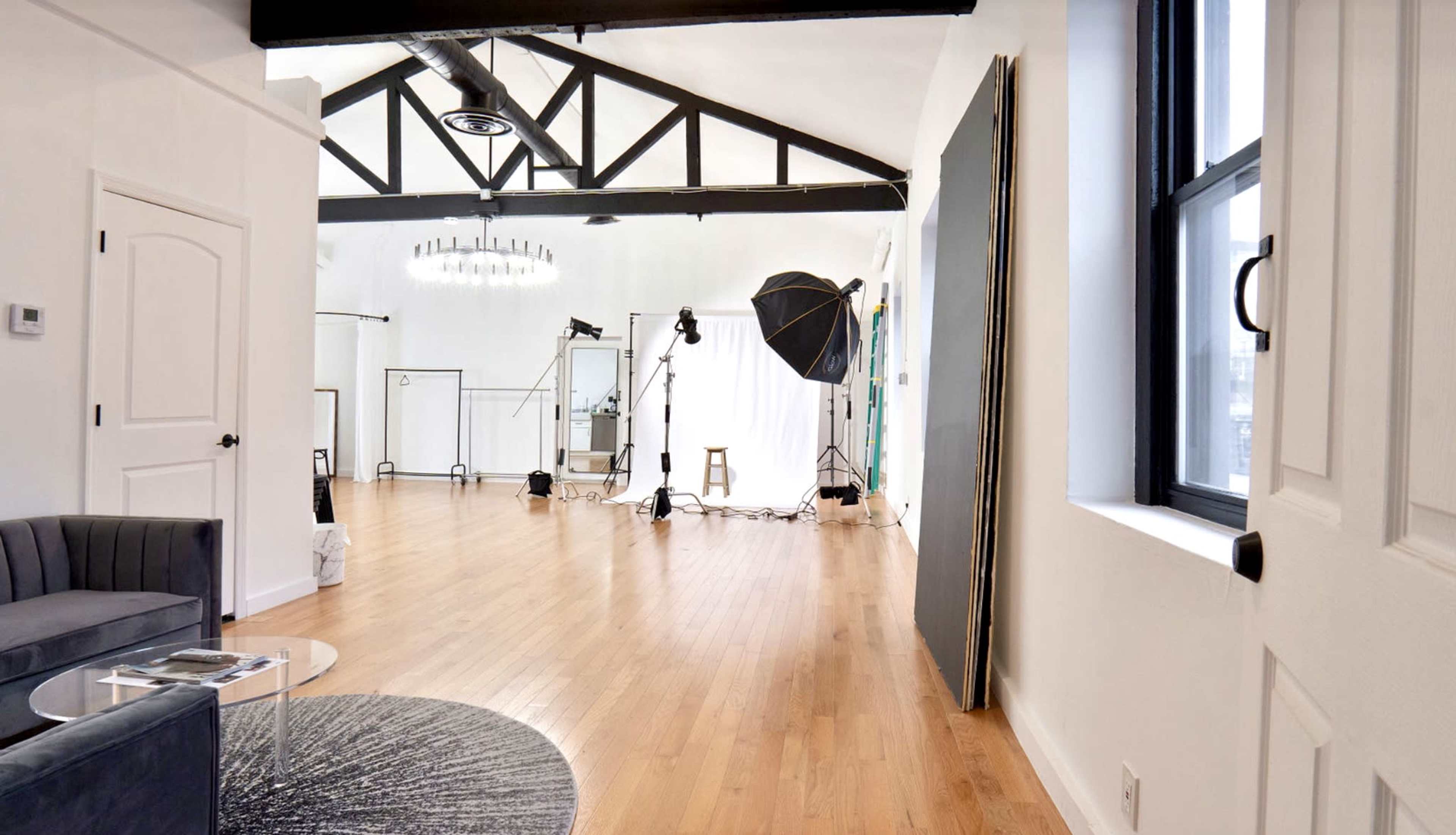 The image shows a spacious, well-lit photography studio featuring hardwood floors, large windows, and various lighting equipment set up for a shoot.