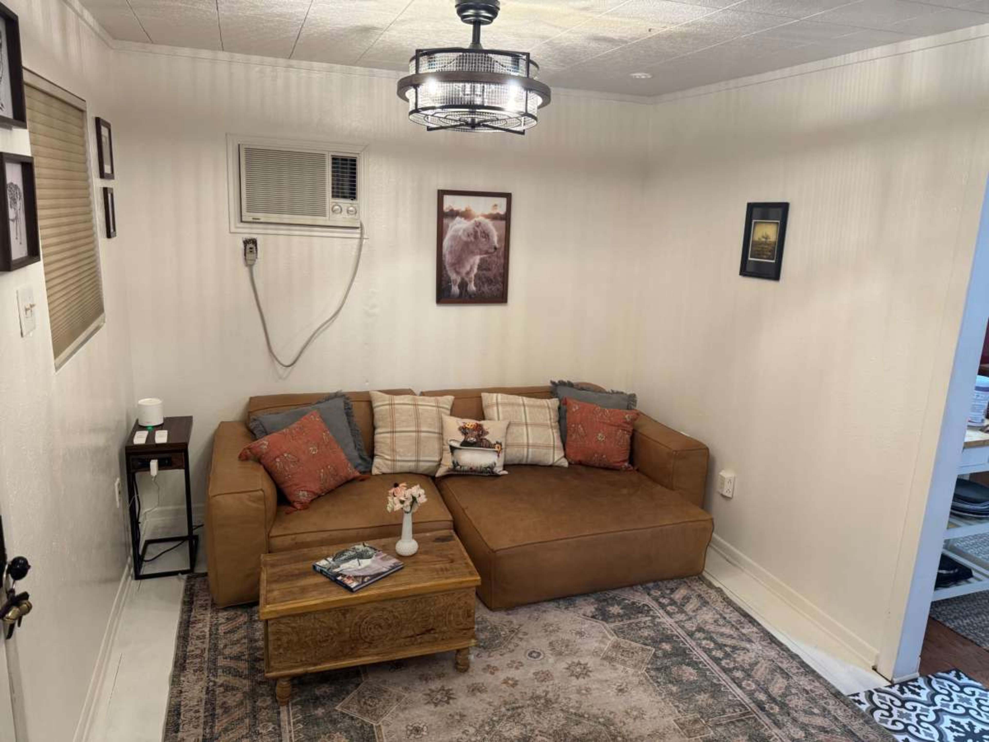 The image shows a small living area featuring a brown sectional sofa with decorative pillows, a wooden coffee table, and an air conditioning unit mounted on the wall.