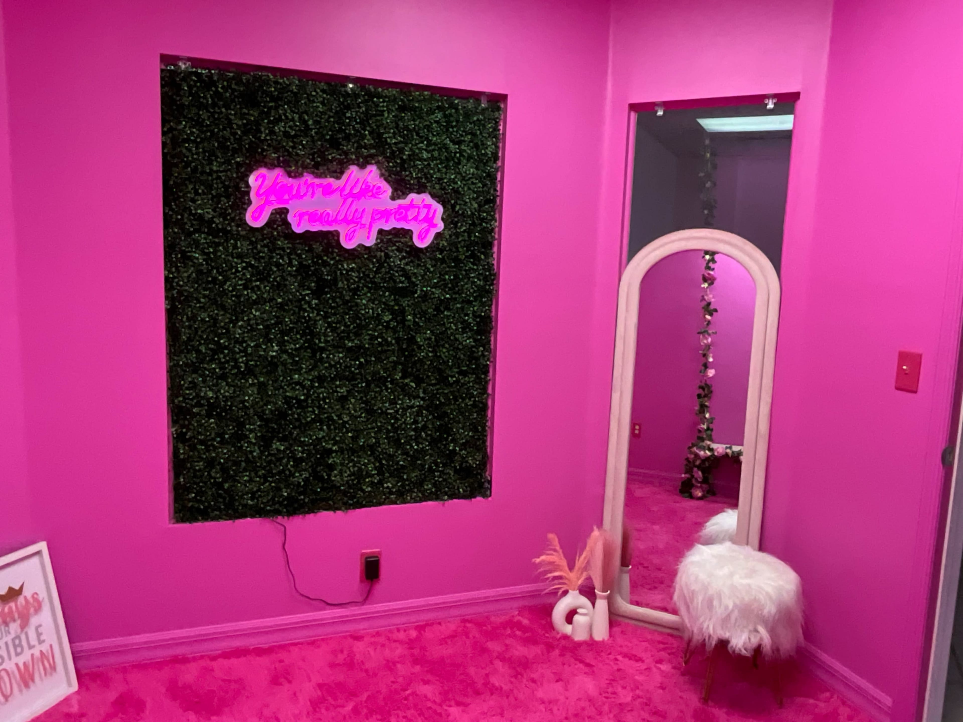 The scene features a vibrant pink room with a neon sign on a green wall, a tall mirror, and a fluffy white chair.
