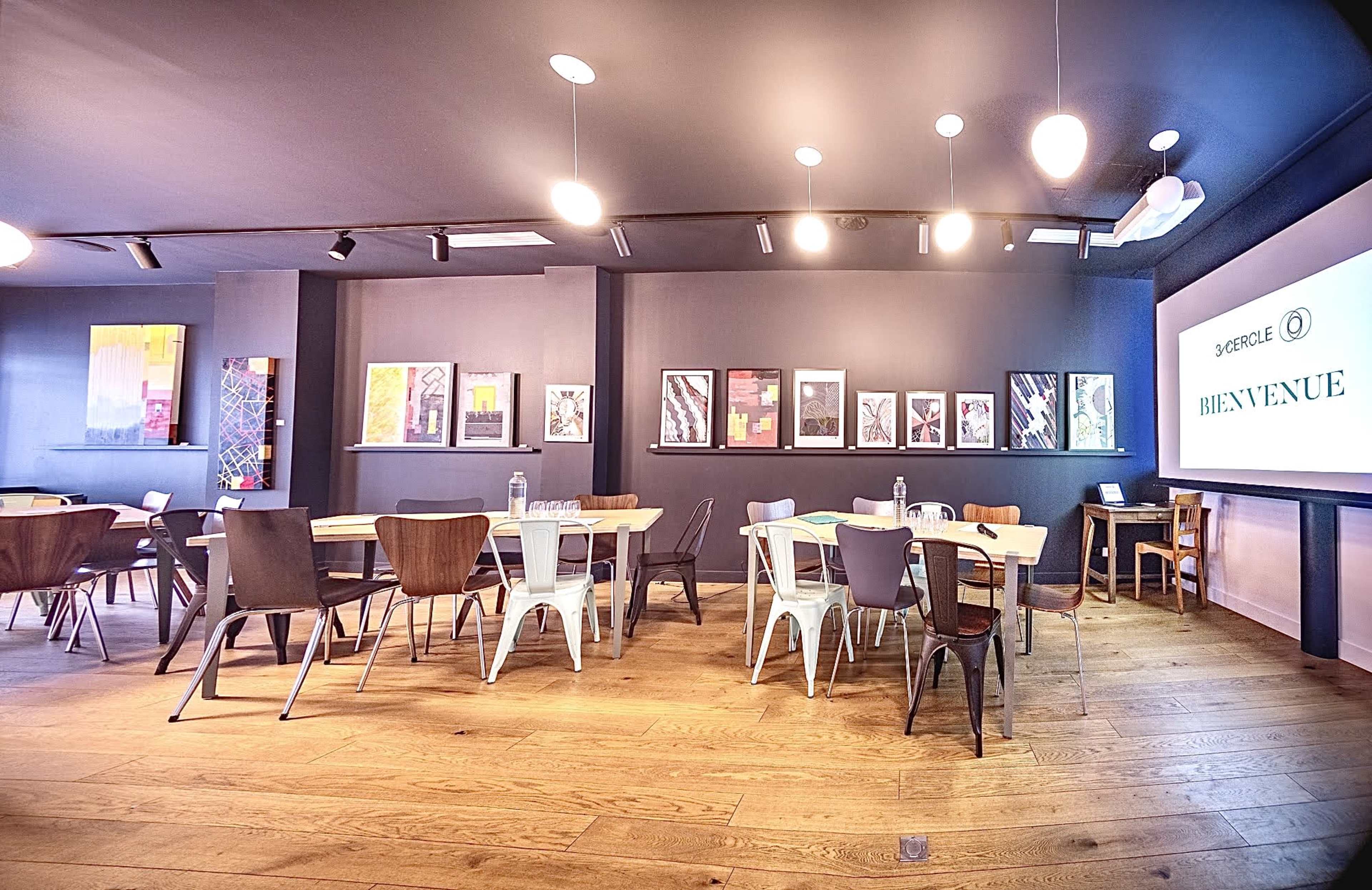 A spacious meeting room features wooden floors, rows of tables and chairs, and framed artwork on the walls.