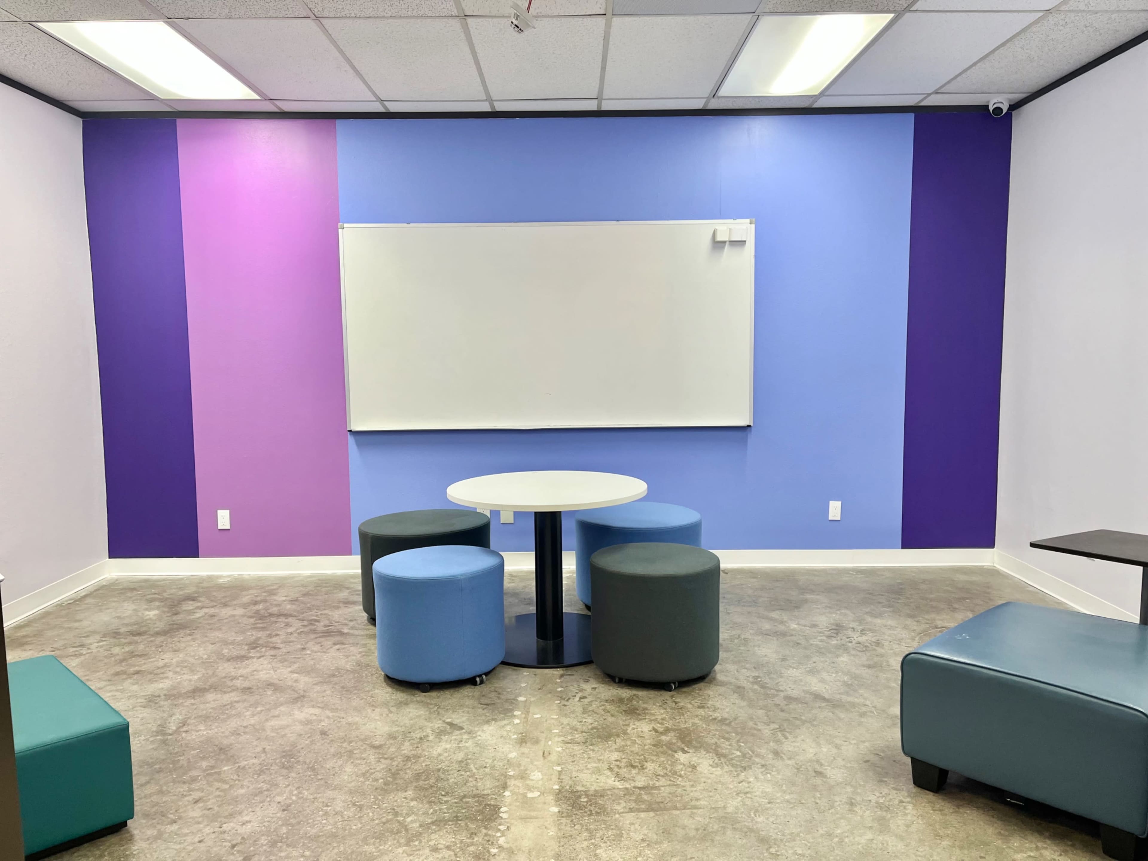 The image shows a small meeting room with a round table and four cylindrical, colorful stools, set against a backdrop of purple and blue walls and a whiteboard.