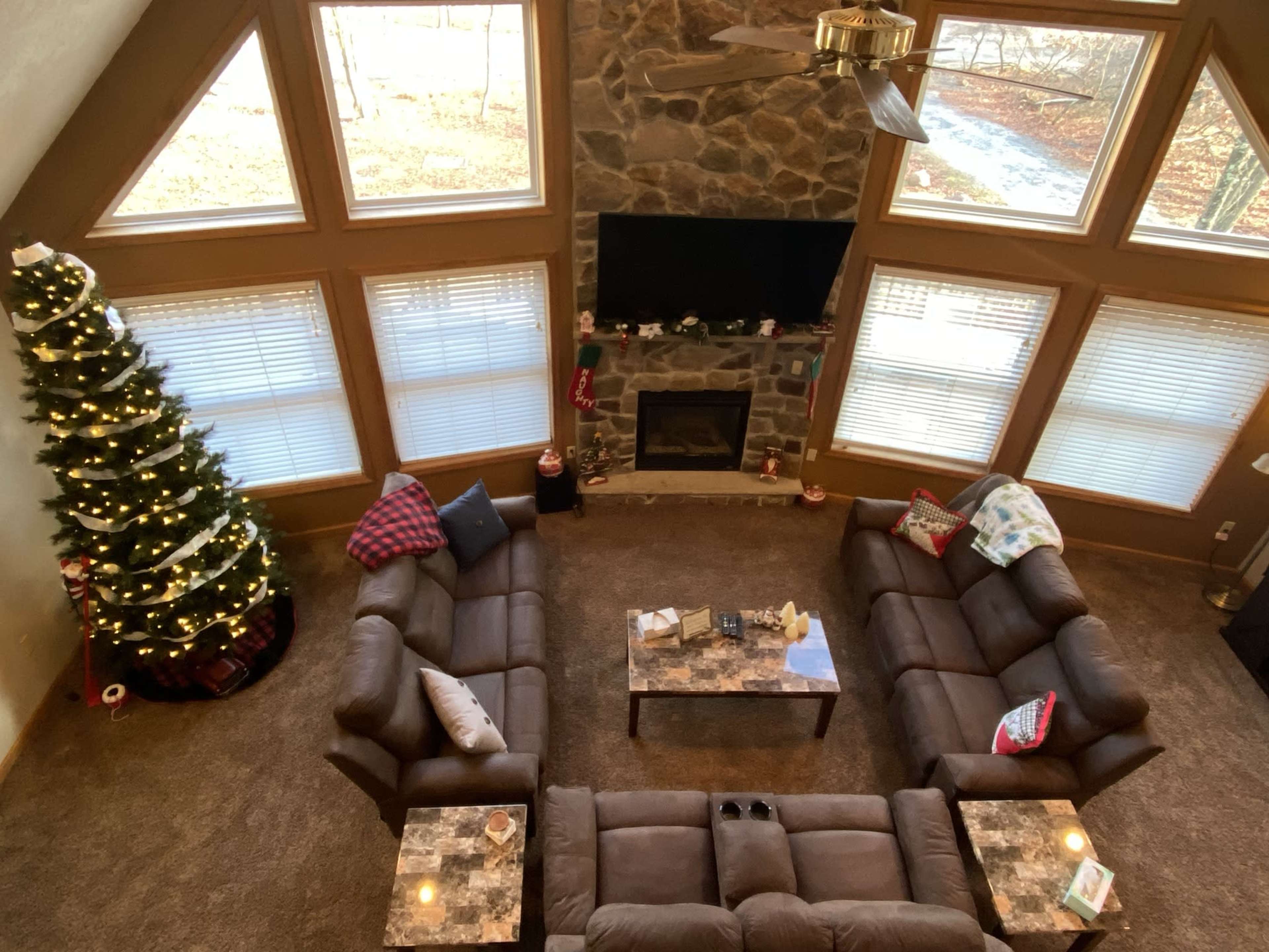 The image shows a spacious living room with a stone fireplace, a large Christmas tree decorated with lights, and two couches arranged around a coffee table.