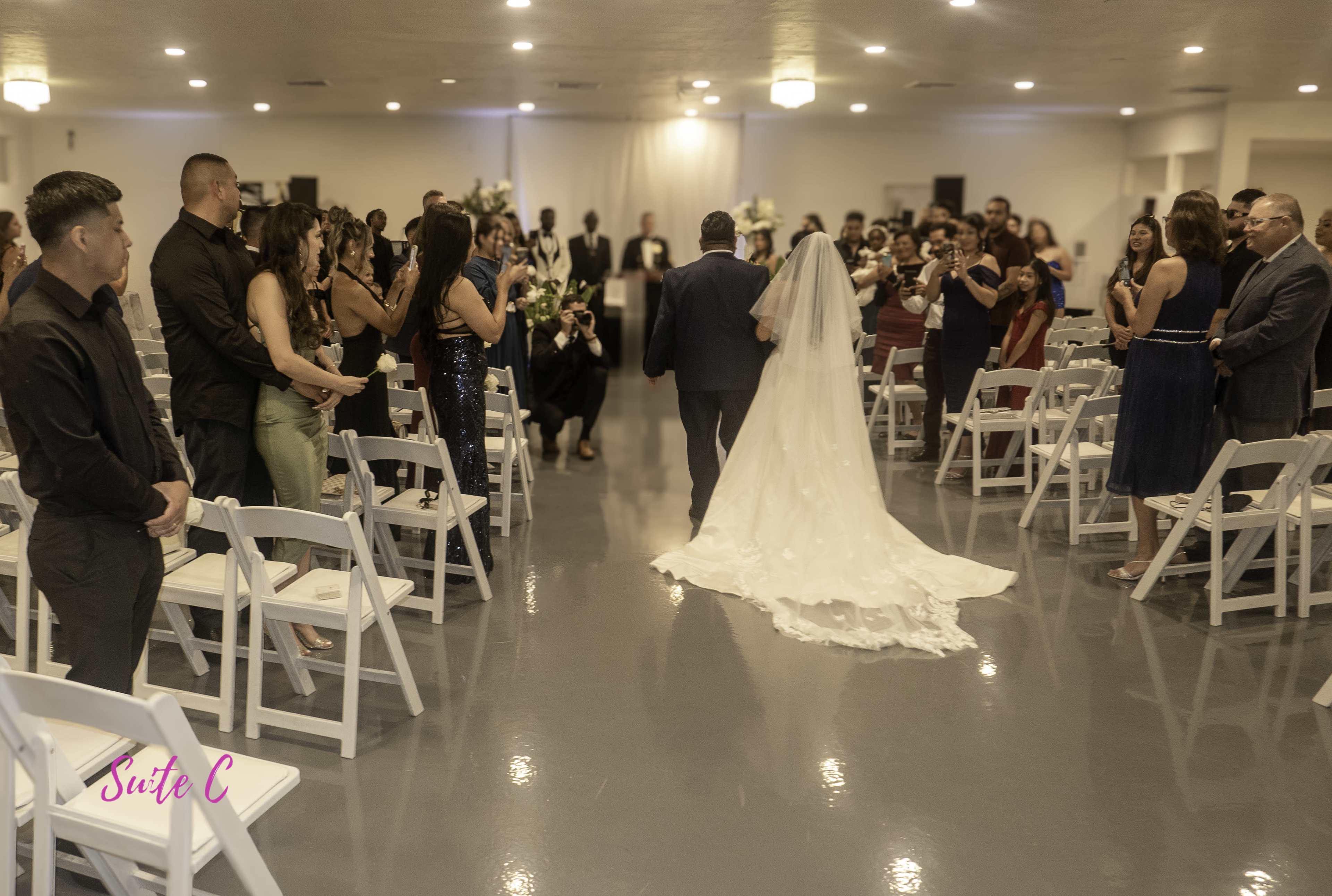 A wedding ceremony is taking place in a venue, with a couple walking down the aisle while guests stand on either side.