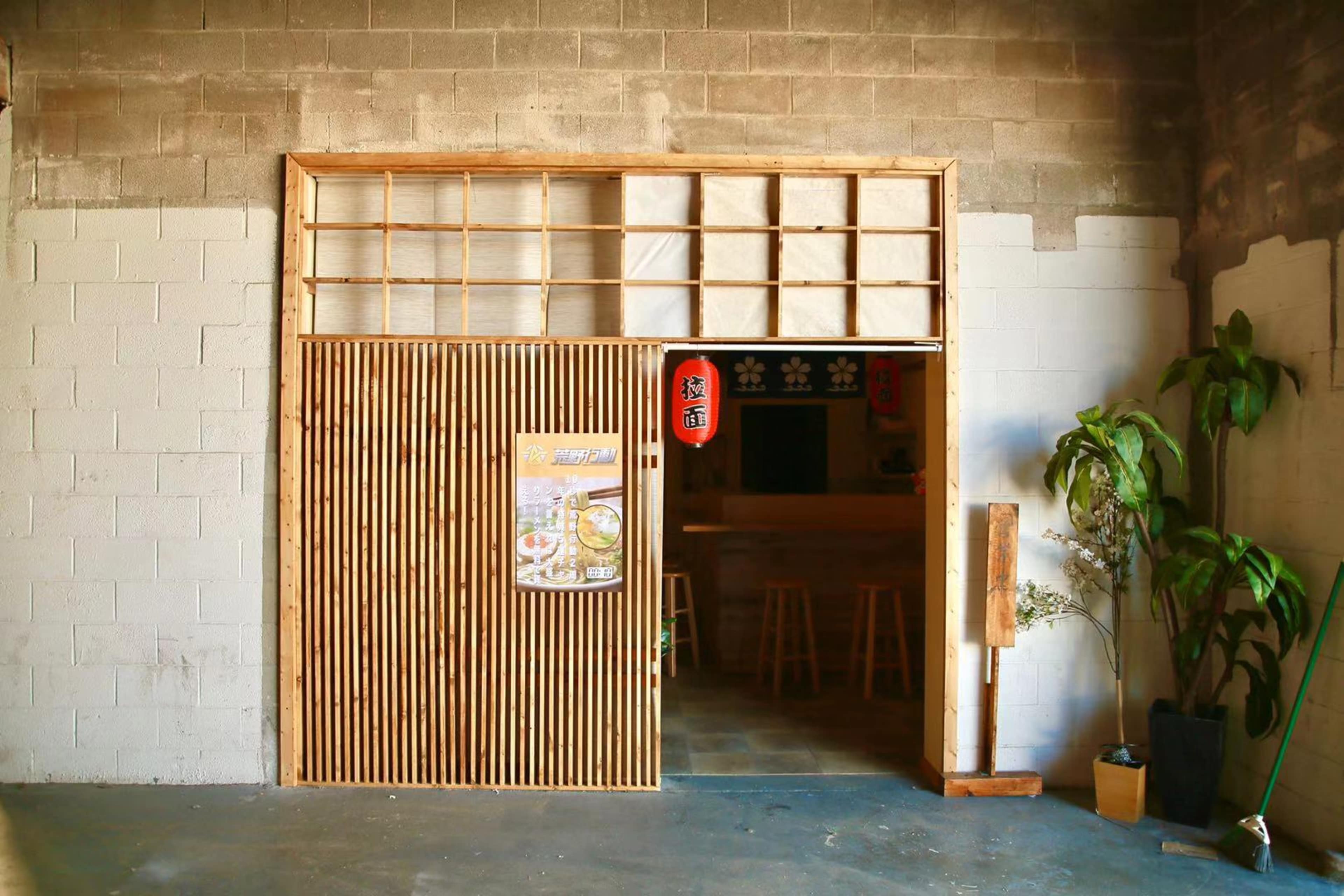 The image shows a doorway framed with wooden slats leading into a minimalist restaurant space with simple seating and decorations.