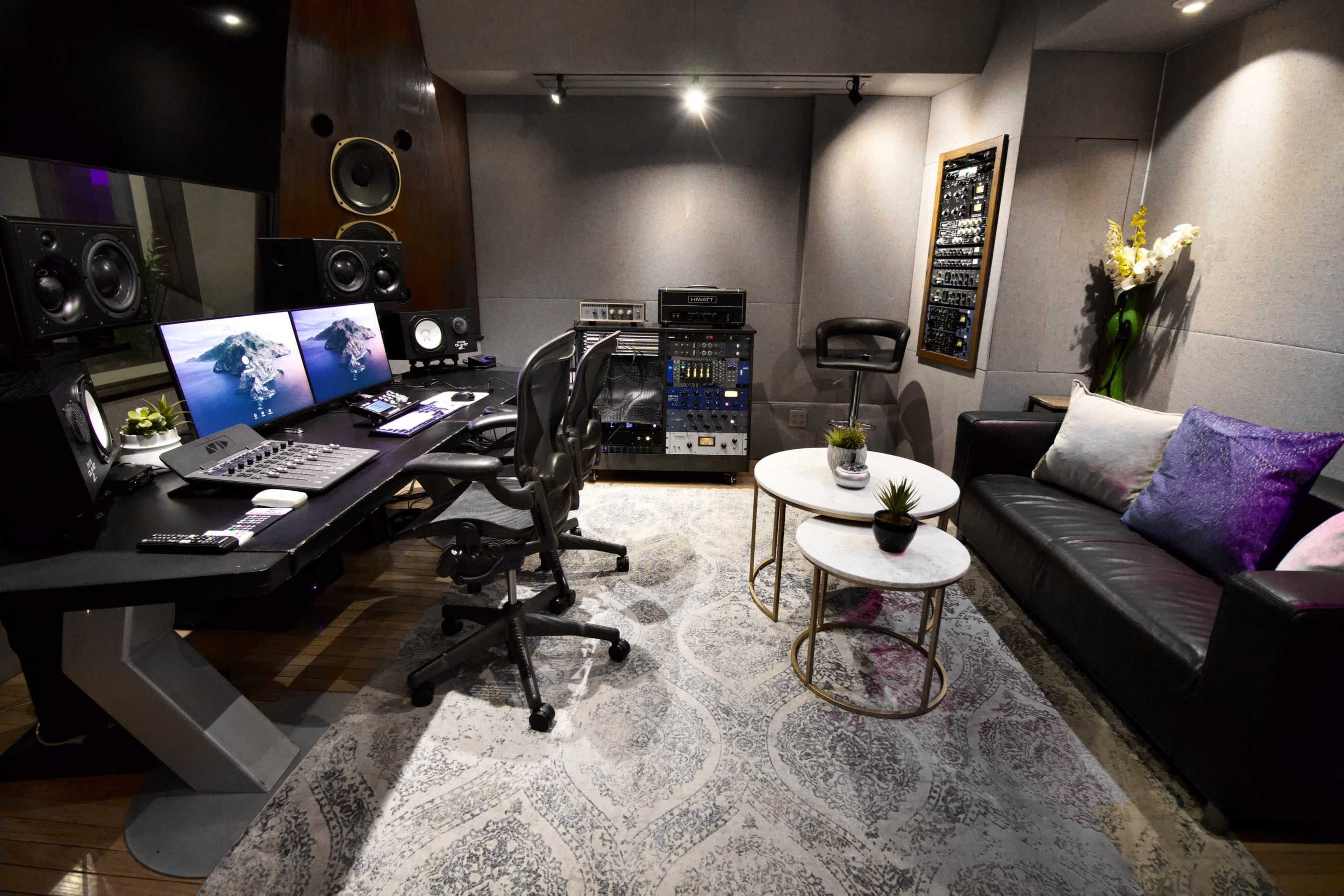 A modern music studio featuring dual computer monitors, a control desk with mixing equipment, a black sofa, and a round coffee table on a patterned rug.